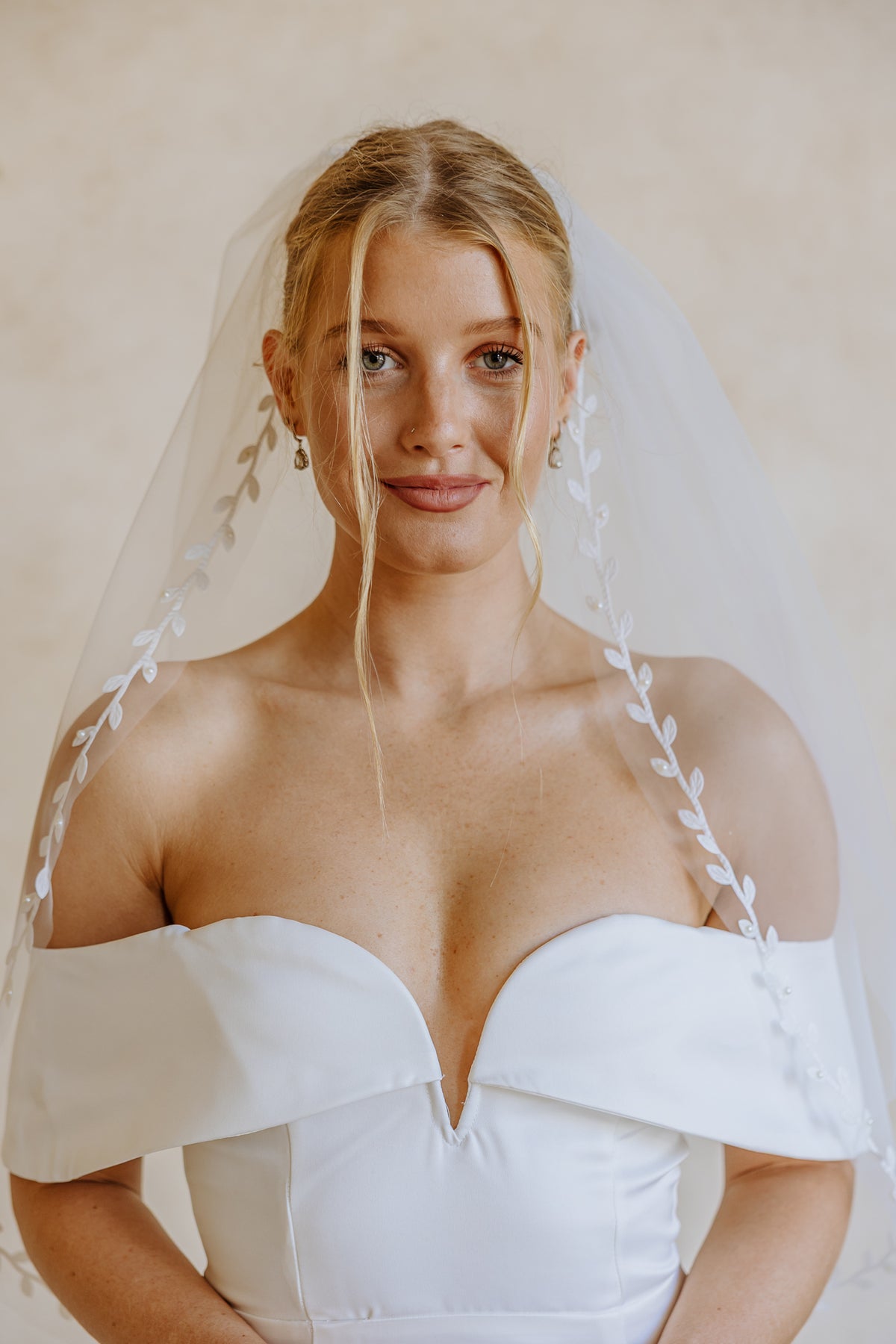 A woman wearing a white off-the-shoulder wedding dress and a sheer white veil with embroidered leaf details, photographed against a soft beige studio background.