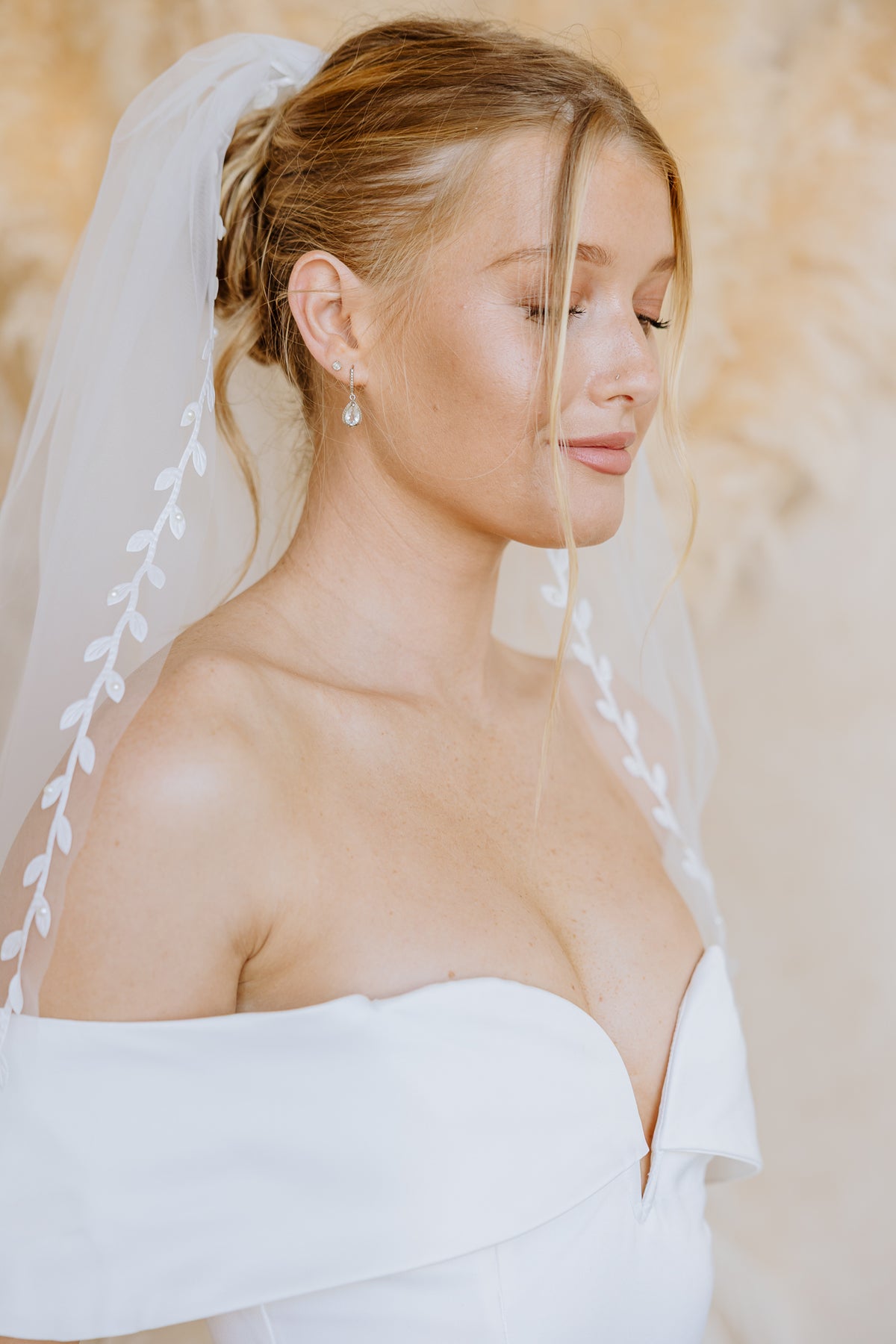 A close-up portrait of a bride wearing a white strapless wedding dress and a sheer veil with embroidered leaf details along the edge. She is shown in profile against a soft, warm neutral background.