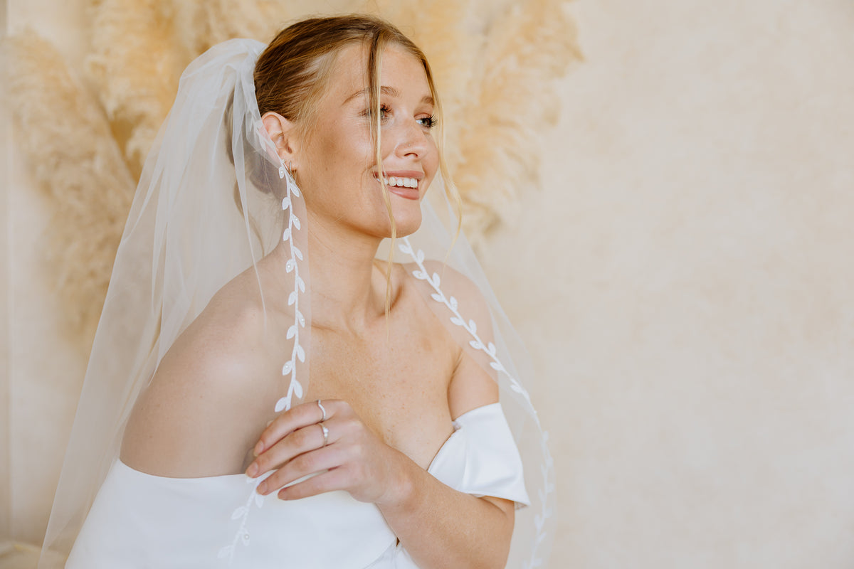 A smiling bride wearing a white off-the-shoulder wedding dress and matching veil with leafy braid details, posed against a warm beige background with dried pampas grass.