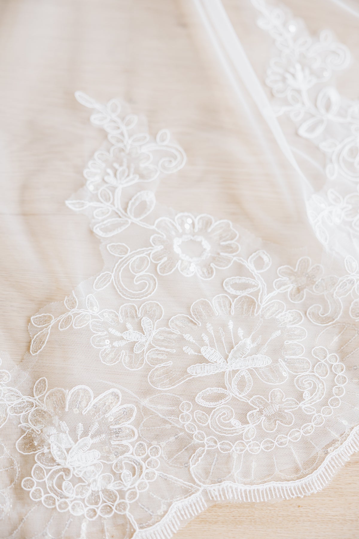 A closeup view of a white floral lace fabric with embroidered flowers and delicate scalloped edges, laid over a soft beige fabric background.