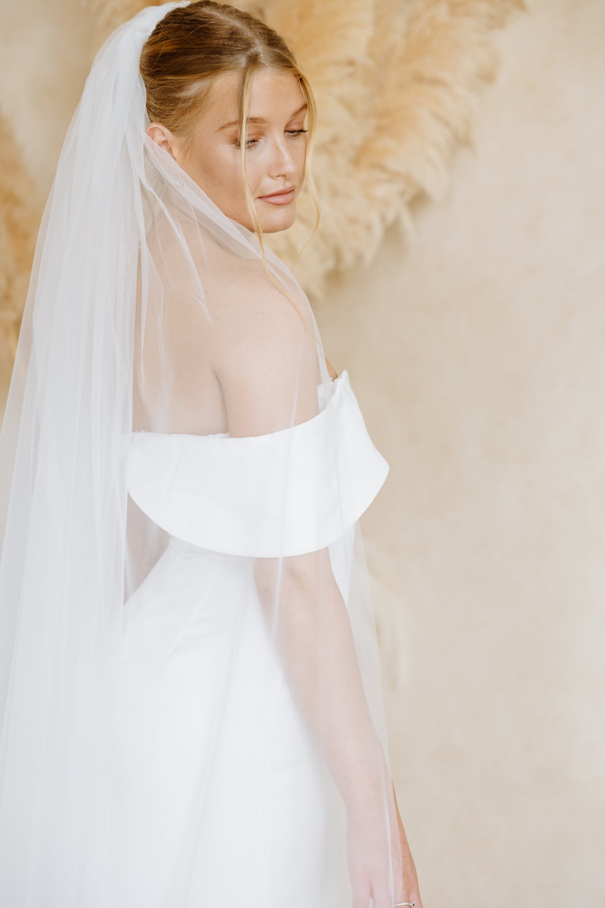A woman wearing a white off-the-shoulder wedding dress with a long sheer veil, posed against a soft beige background with a decorative feather wall hanging.