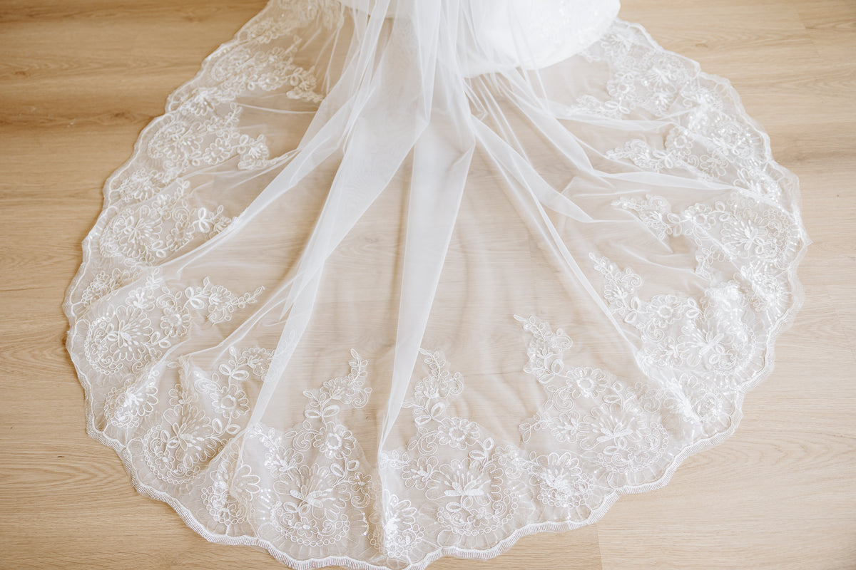 White bridal veil with intricate floral lace and tulle layers, spread out on a light wood floor.
