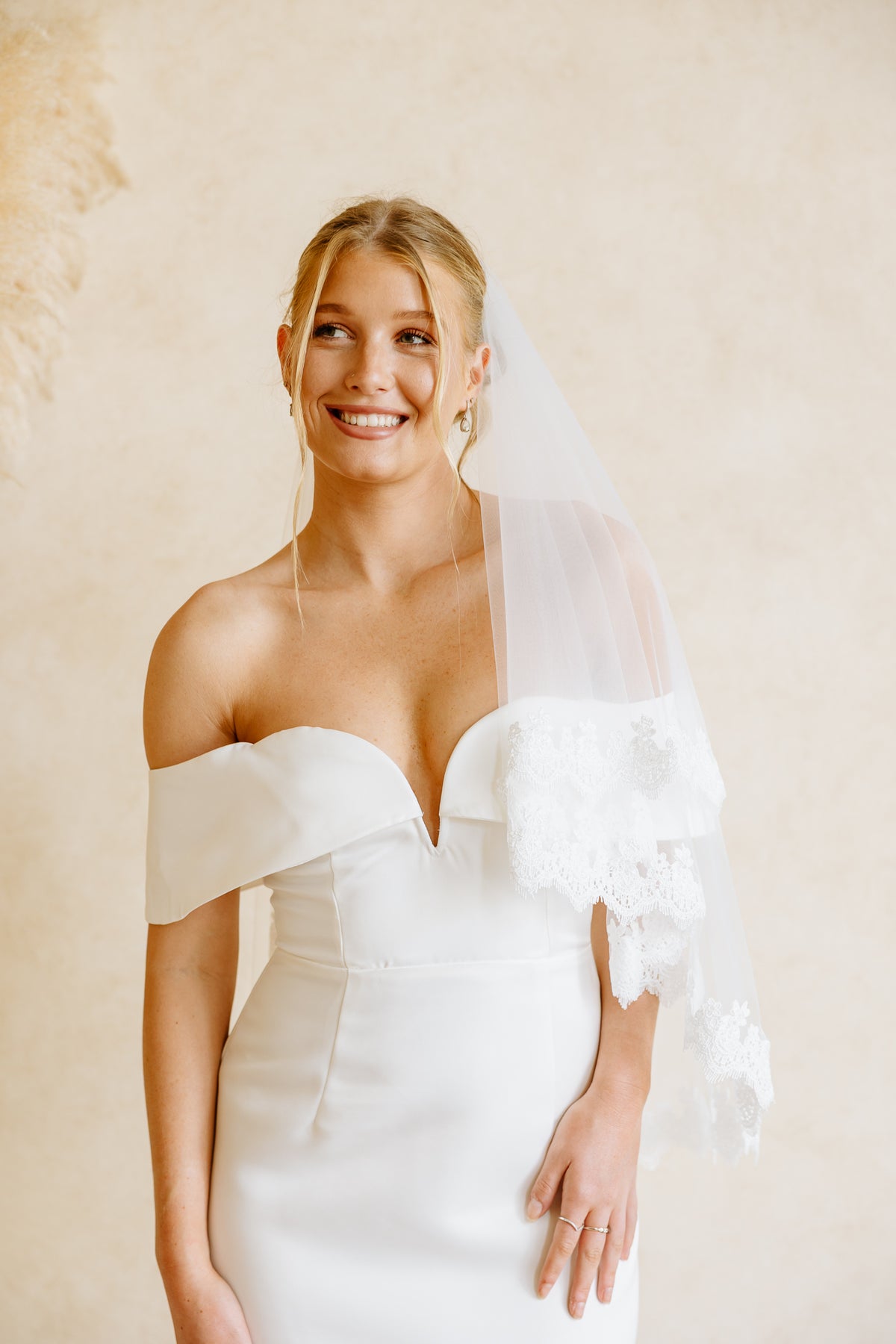 A smiling blonde bride wearing an off-the-shoulder white wedding dress with a lace-trimmed sheer veil, posed against a warm beige textured wall.