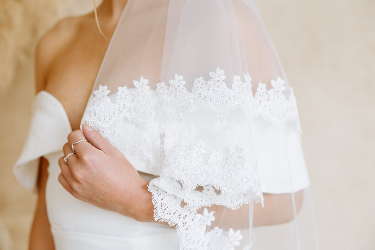 A close-up of a bride wearing a white off-the-shoulder wedding dress with a wedding veil that has intricate floral lace trim draped over her shoulder.