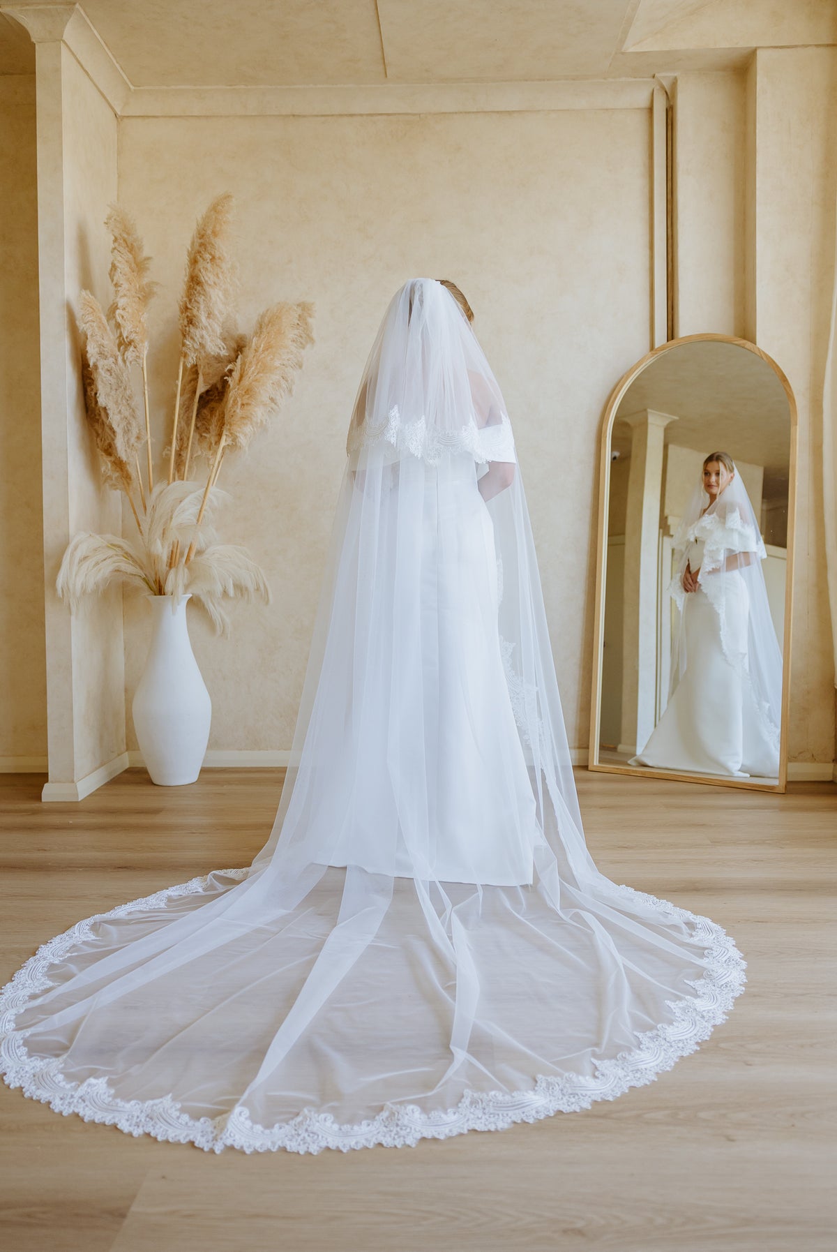 A bride in a white wedding gown and long sheer veil stands with her back to the camera in a warm beige room, with a large mirror showing her front reflection and dried grass decor in a white vase.