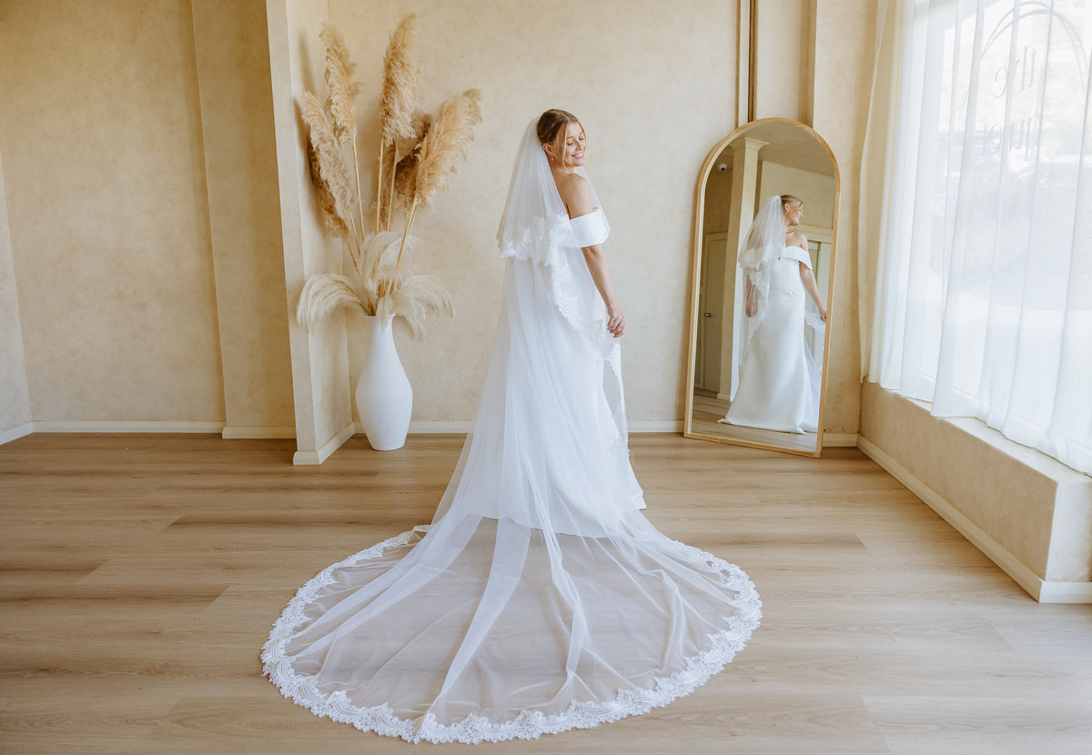 A bride in a white off-the-shoulder wedding dress with a long lace-trimmed veil stands in a bright room with wood floors, a large arched mirror, and pampas grass in a vase.