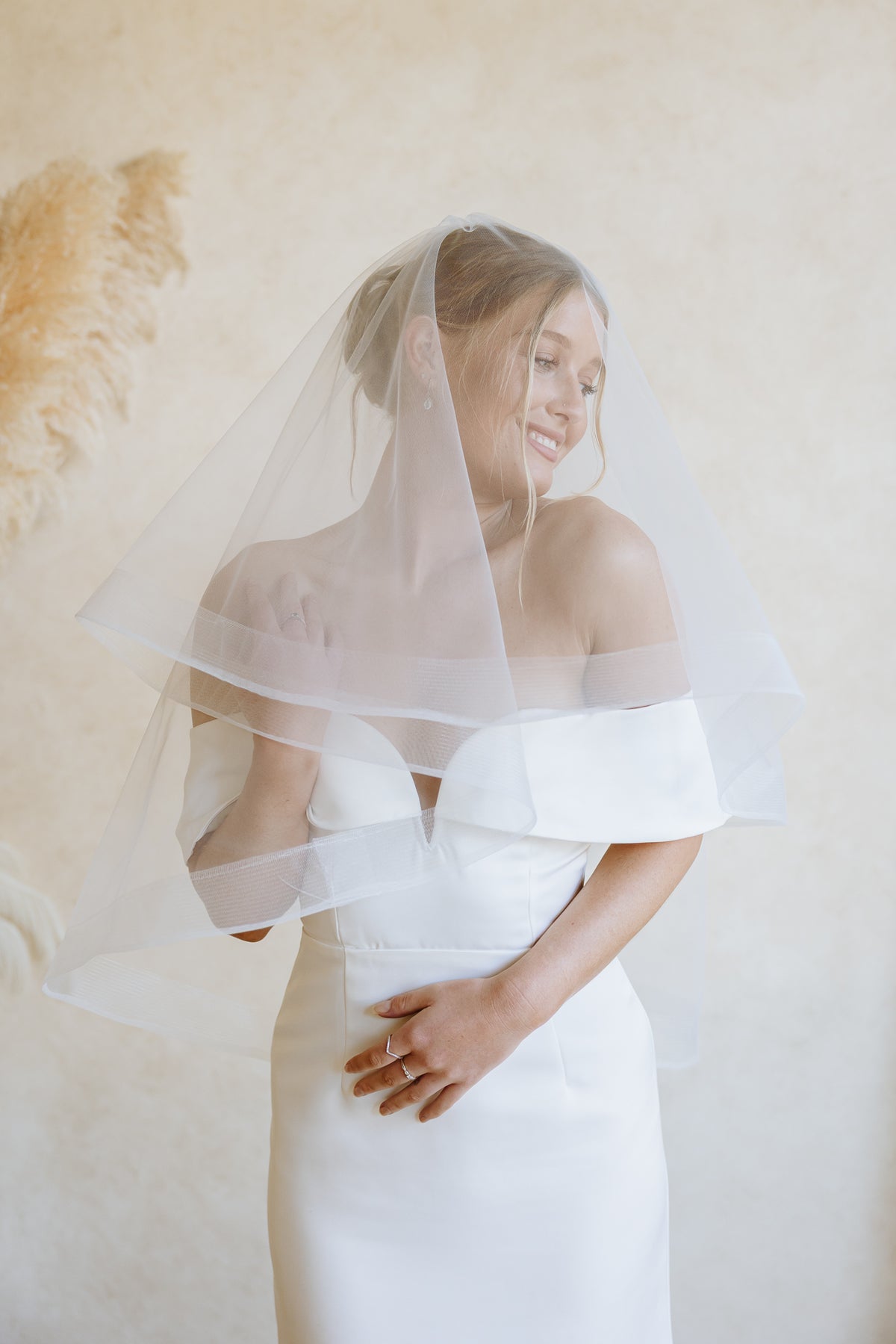 A woman wearing a white off-the-shoulder wedding dress and a sheer white blusher veil with horsehair trim, posed against a warm beige textured wall with dried pampas grass decor.