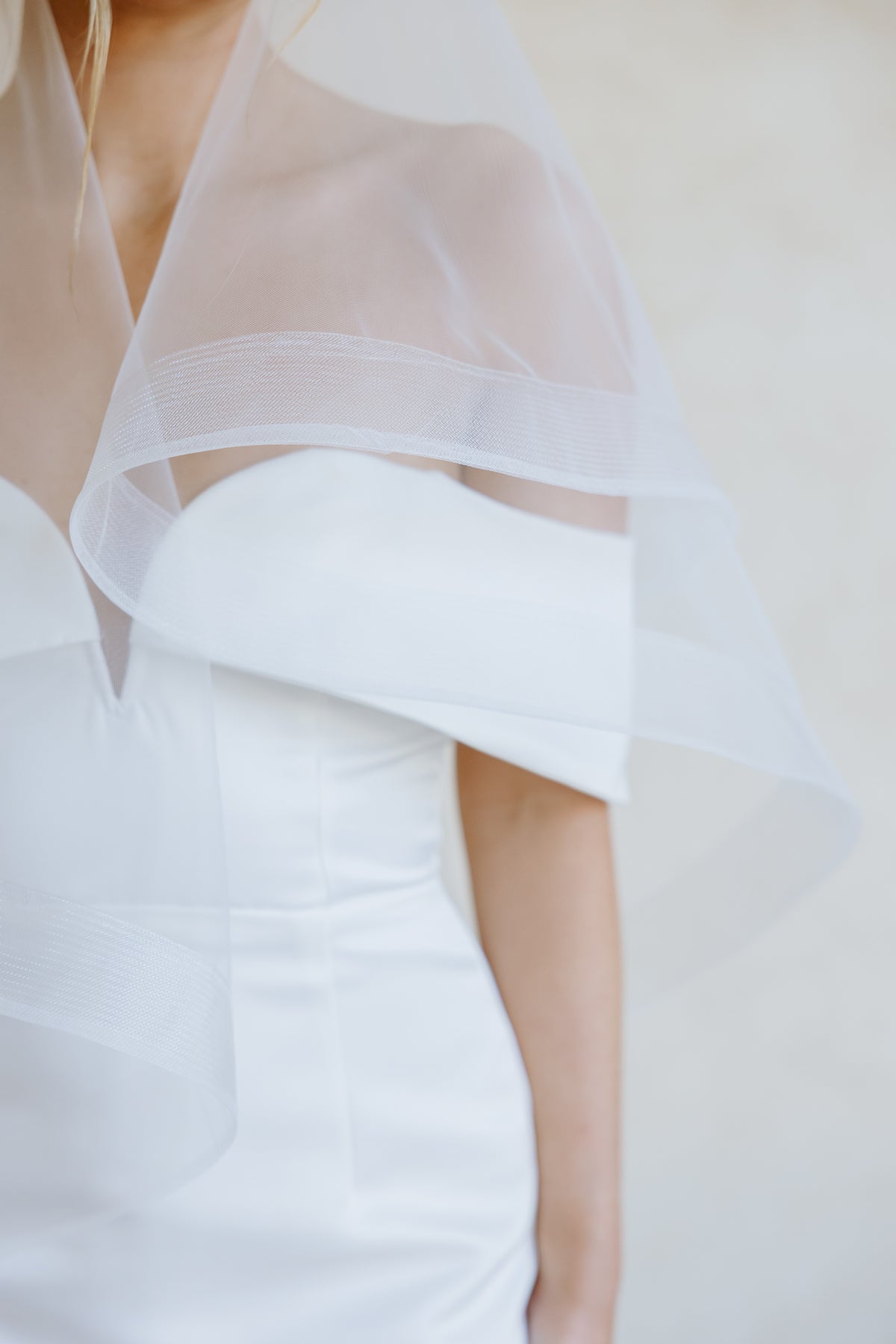 Close-up of a woman wearing a white off-the-shoulder wedding dress with a sheer white veil with horsehair trim draped over her shoulder, against a soft light beige background.