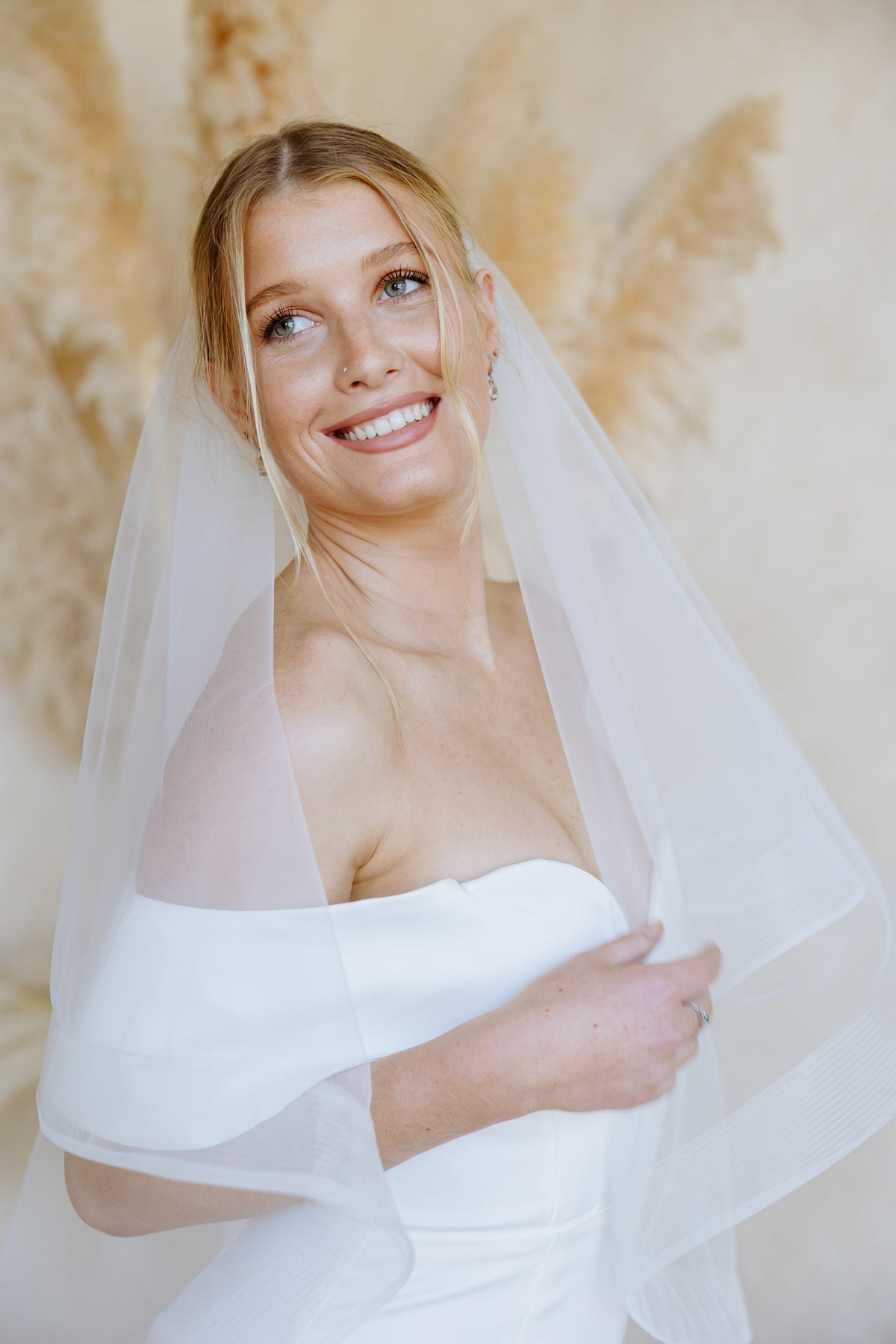 A smiling blonde bride wearing a white off-the-shoulder wedding dress and a sheer horsehair trim veil, posed against a warm beige background with pampas grass.