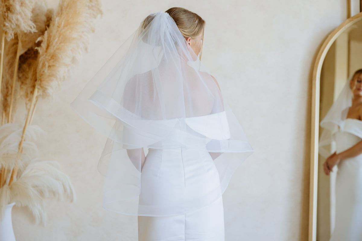 A woman wearing a white off-the-shoulder dress and a long sheer tulle bridal veil, shown from the back in a softly lit indoor setting with a mirror and pampas grass decor.