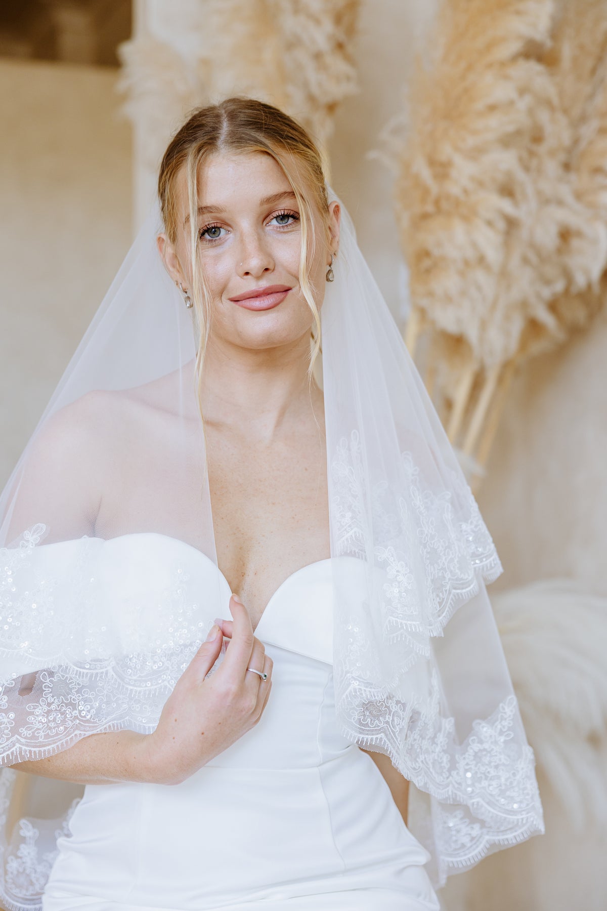 A blonde bride wearing a white strapless wedding dress with lace ruffle sleeves and a matching sheer lace-trimmed veil, posed against a soft neutral background with dried pampas grass.
