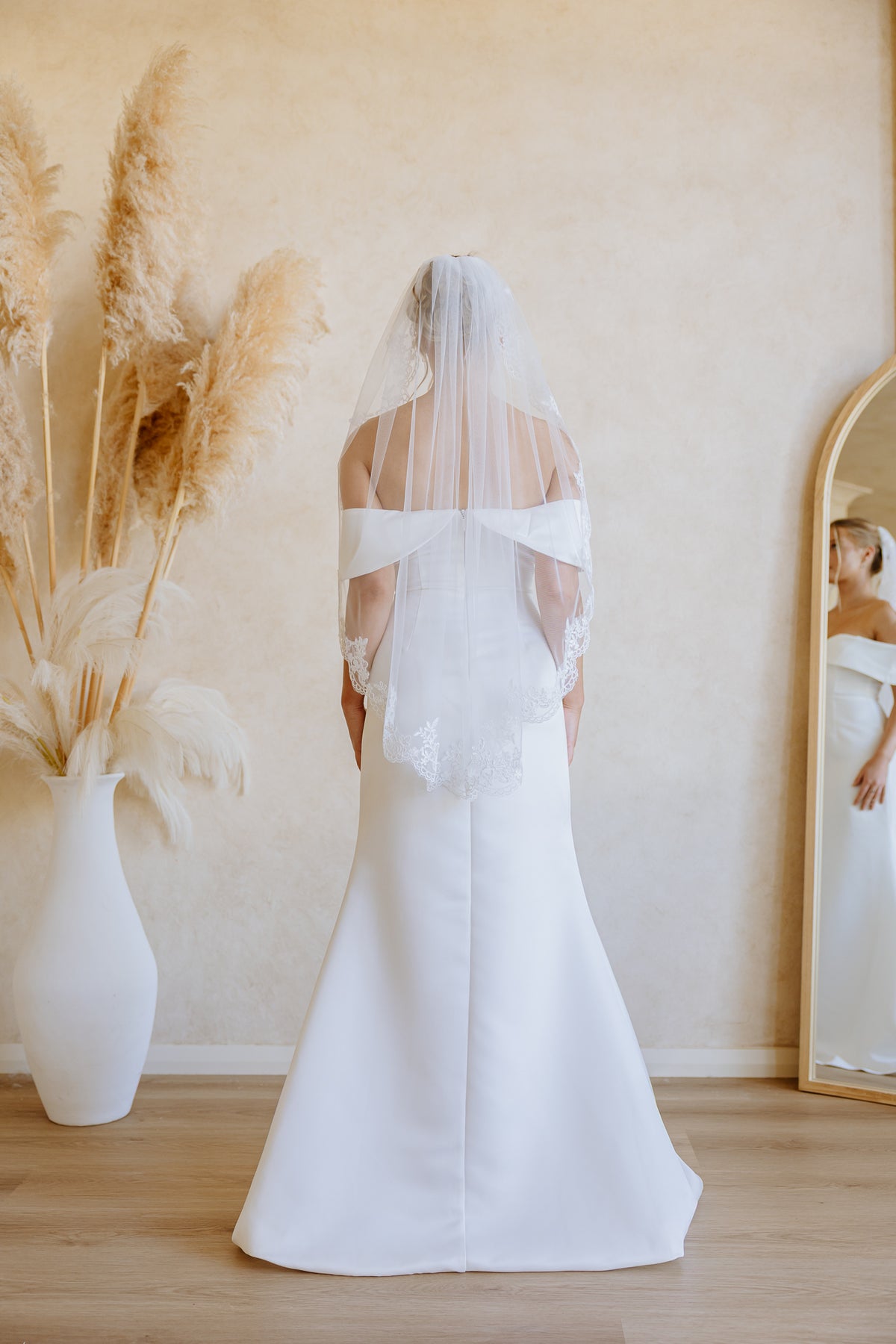A woman wearing a white off-the-shoulder wedding dress with a long sheer veil, standing with her back to the camera in a softly lit room with a mirror, pampas grass in a vase, and light wood flooring.