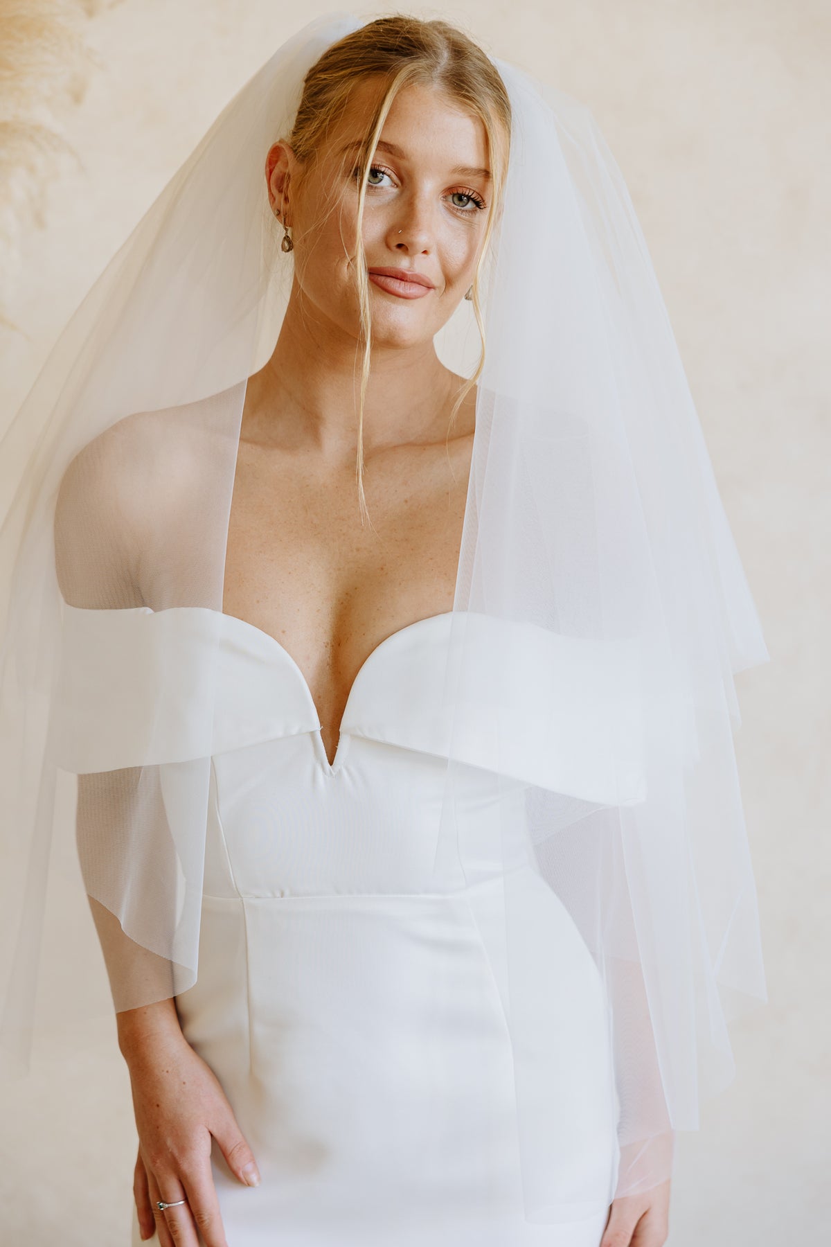 A bride wearing a white off-the-shoulder wedding dress with a layered tulle veil, photographed against a soft beige background.