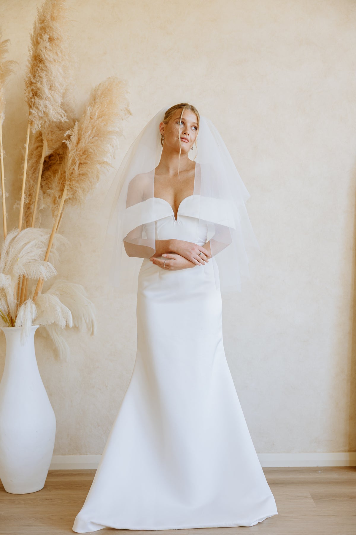 A woman wearing a white off-the-shoulder wedding dress with a sweetheart neckline, paired with a soft tulle veil that flowy, standing in a warm beige room with pampas grass in a white vase.