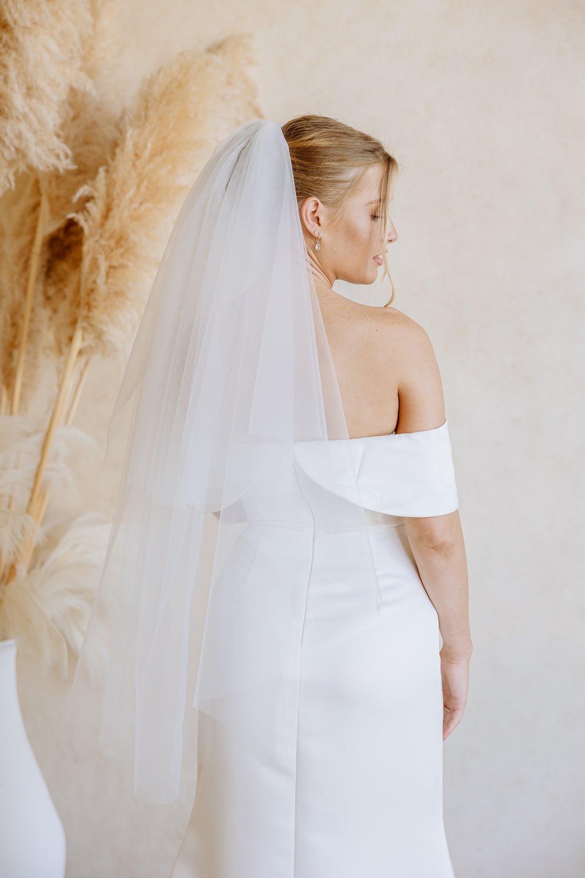 A woman wearing a white off-the-shoulder wedding dress and a long sheer tulle veil, photographed from the back against a warm beige wall with pampas grass decor.