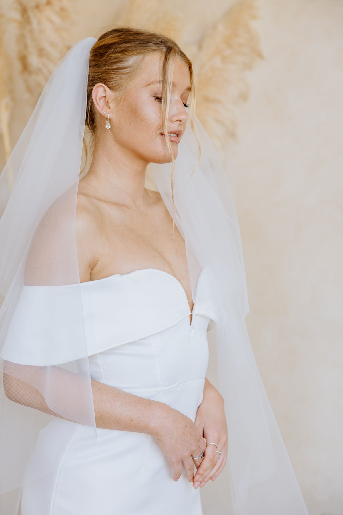 A woman wearing a white off-the-shoulder wedding dress with a sheer veil, posed against a soft beige background with dried floral decor.