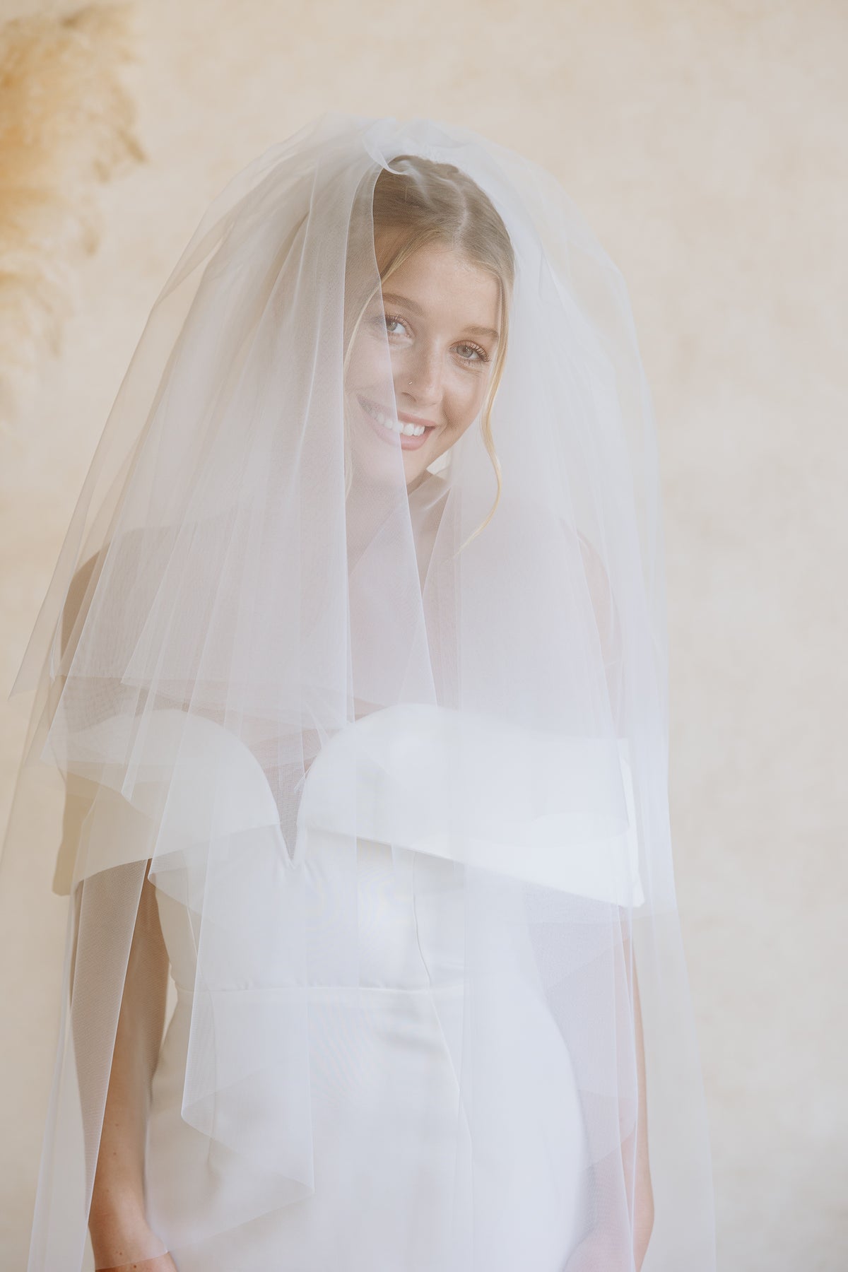 A smiling bride wearing a long, flowing white tulle bridal veil over a white wedding dress, posed against a soft beige textured wall.