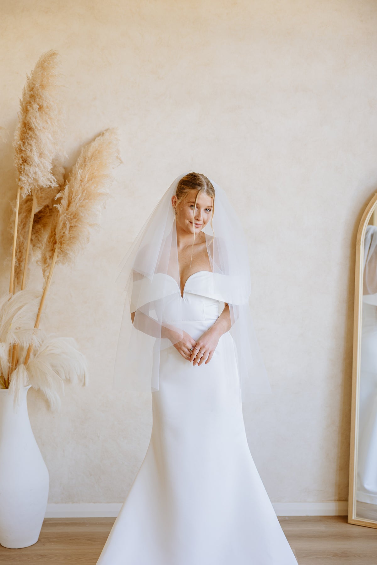 A woman wearing a white wedding dress with a sheer veil stands indoors against a warm beige wall, with dried pampas grass in a white vase on the left and part of a mirror on the right.