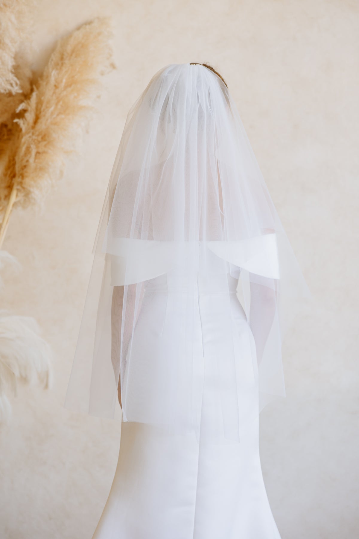 A white bridal veil with a wide matching band, shown from the back on a model wearing a white wedding dress against a warm beige background with dried pampas grass.