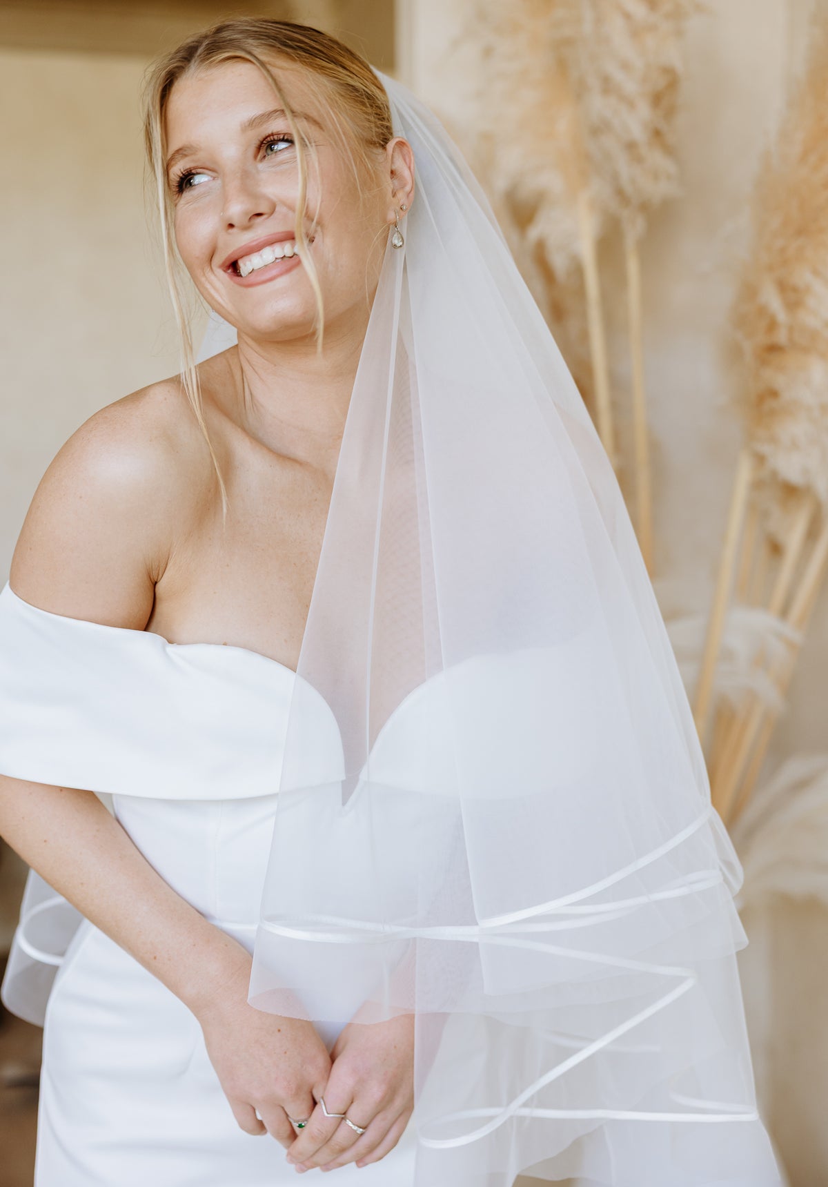 A smiling bride with blonde hair wears a white off-the-shoulder wedding dress and a sheer veil with a ribbon edge, standing near dried pampas grass.