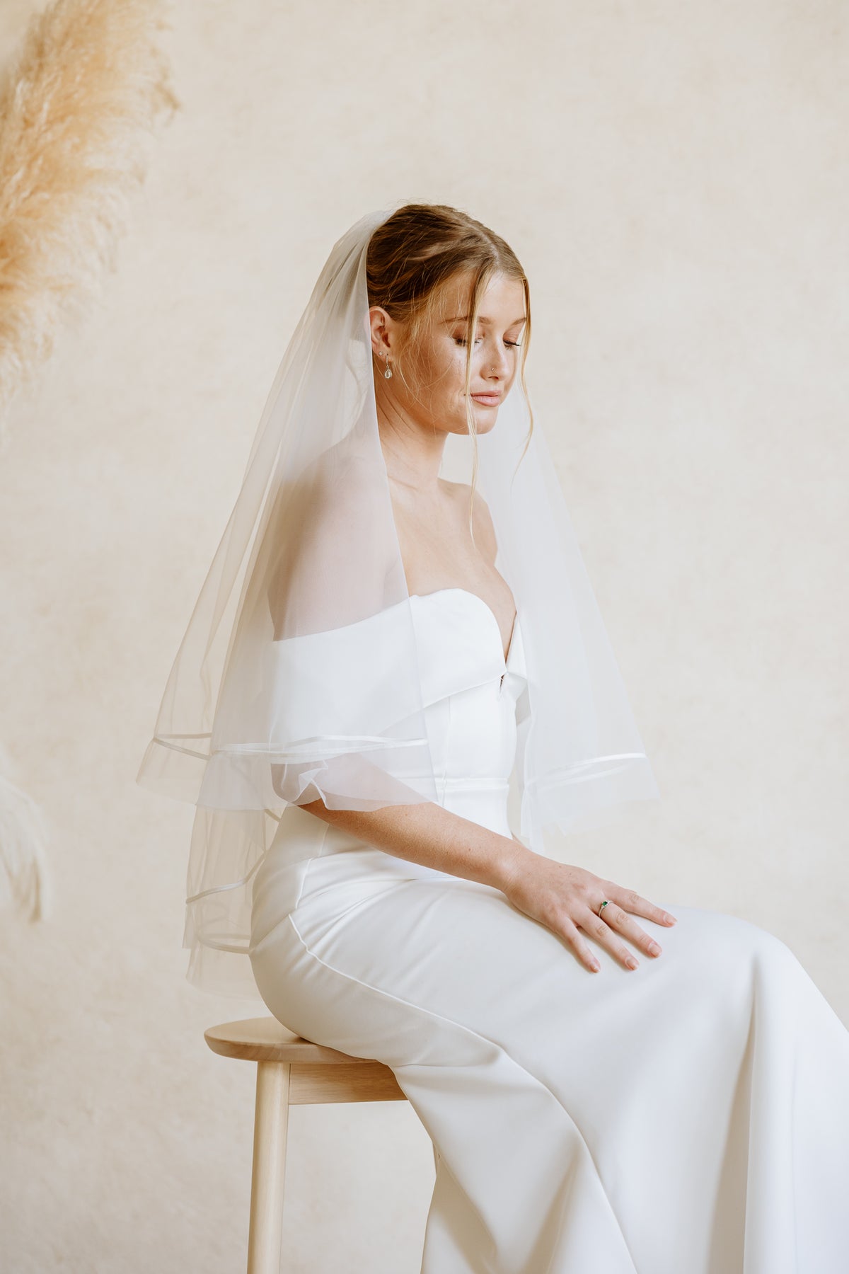 A bride is wearing a off-shoulder wedding dress in white and a sheer white bridal veil with ribbon trim, while sitting on a wooden stool