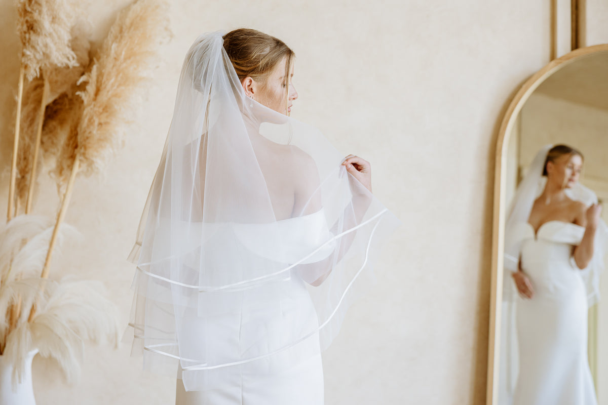 A bride in a white off-the-shoulder dress and veil looks into an arched mirror, with tall pampas grass stems visible in the background of the room.