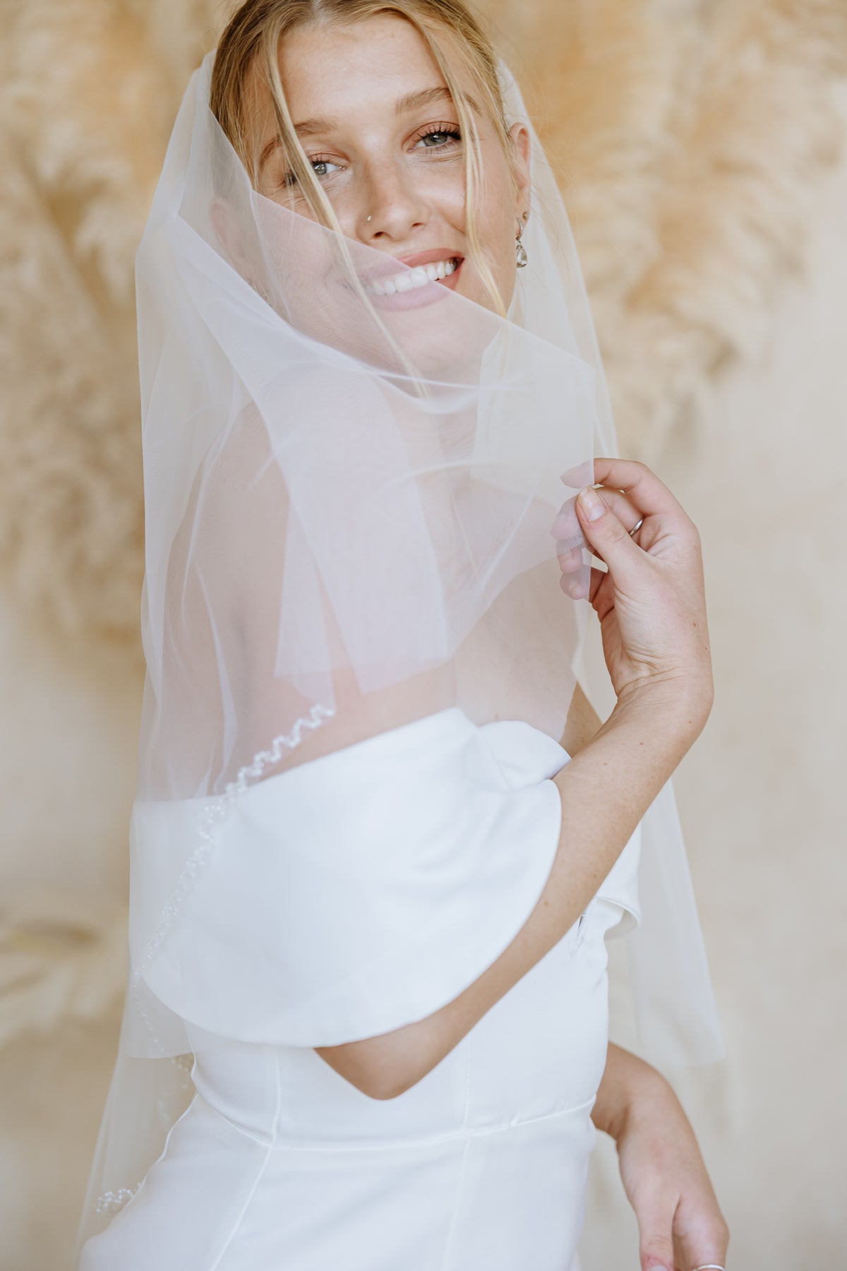 A smiling bride wearing a sheer white bridal veil and a white wedding dress, holding the veil in front of her face against a soft beige textured background.