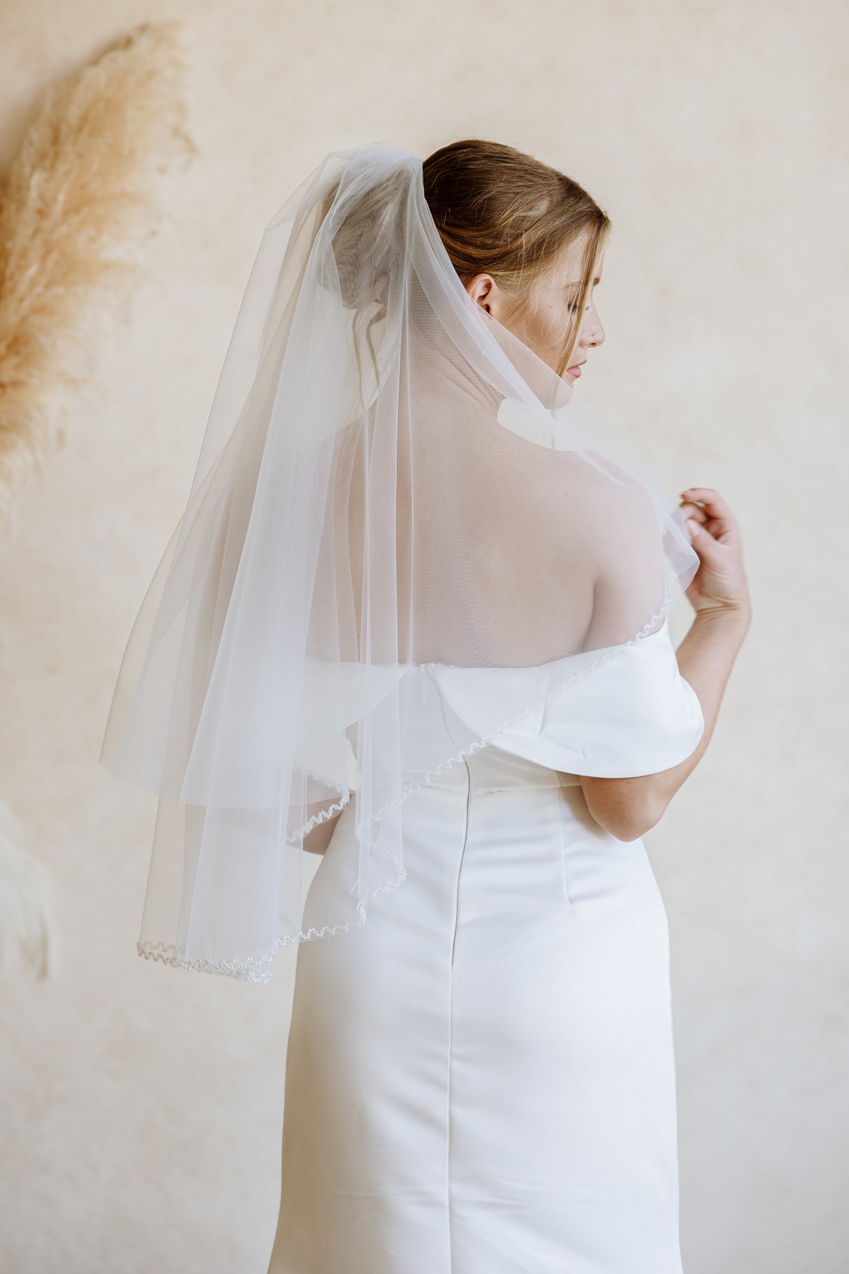 A woman wearing a white wedding dress and a long sheer tulle bridal veil, shown from the back and side against a soft beige wall with dried pampas grass decor.