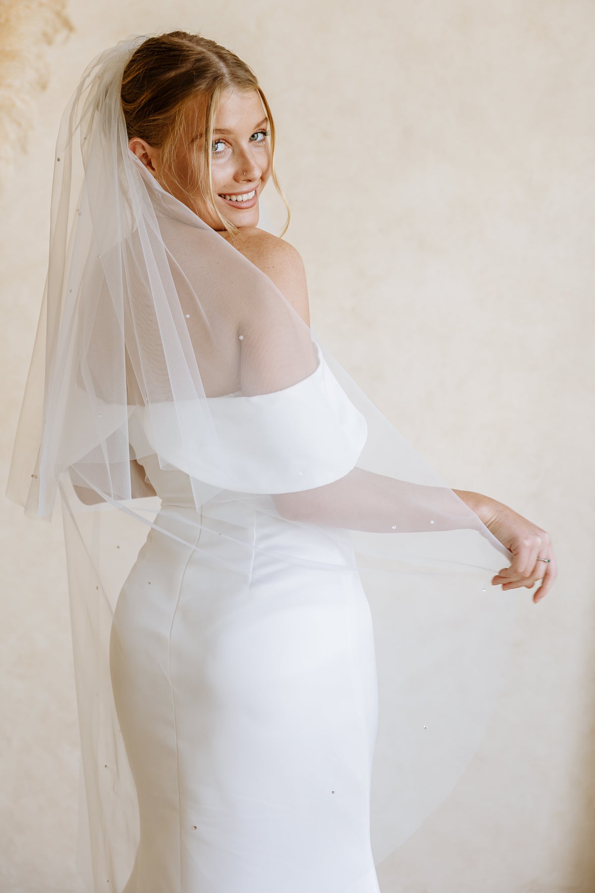 A smiling bride in a fitted white wedding dress with a sheer off-the-shoulder veil, posed against a soft beige textured wall.