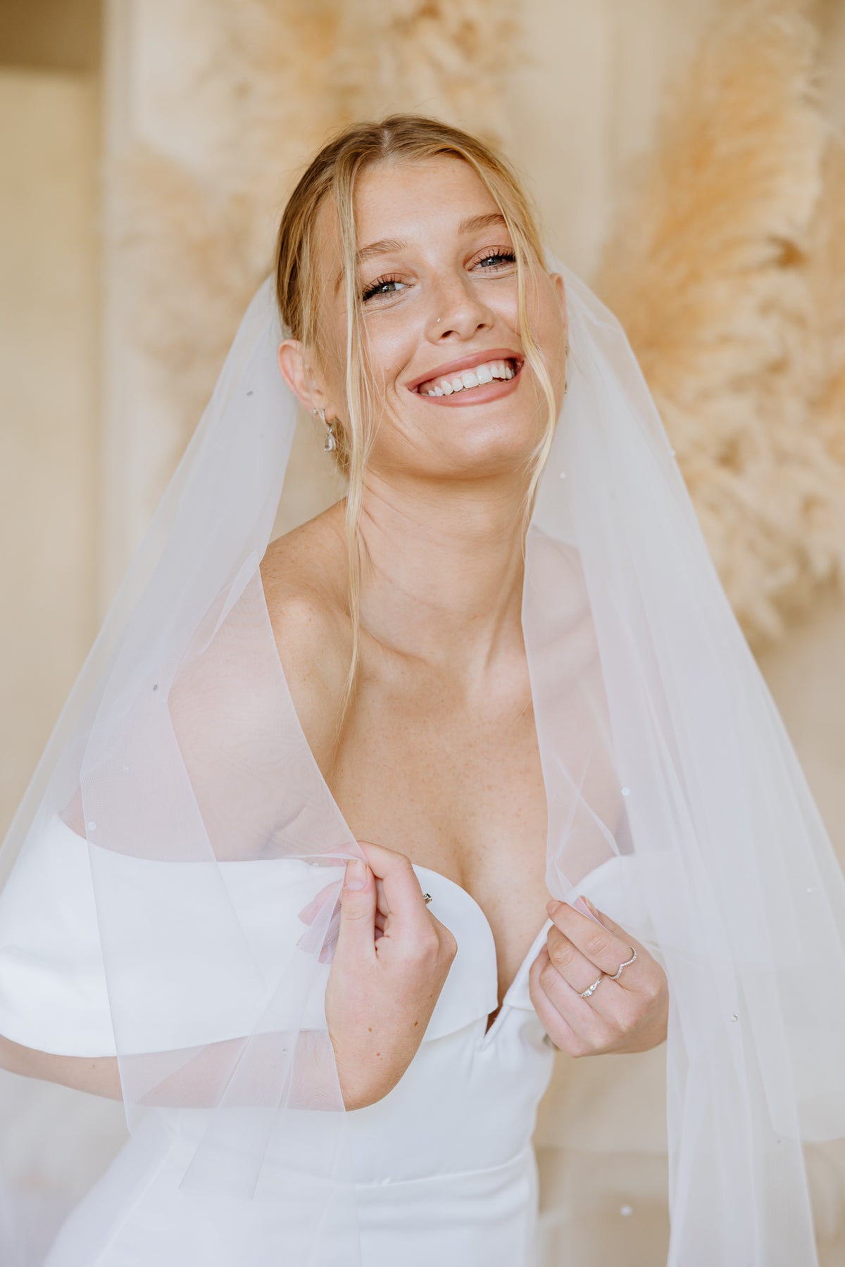 A smiling bride wearing a white wedding dress and long sheer veil, posed indoors against a soft neutral background with dried floral decor.