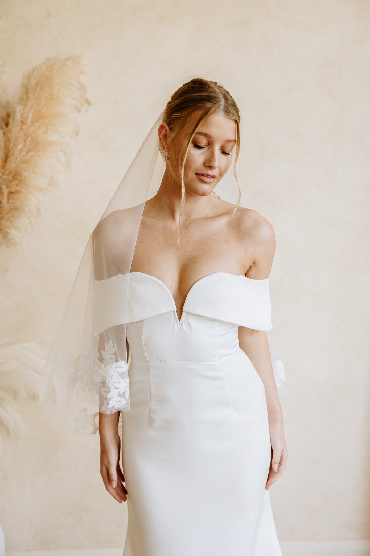 A woman wearing a white off-the-shoulder wedding dress with a sweetheart neckline, lace-trimmed sleeves, and a sheer veil, standing against a warm beige wall with pampas grass decor.