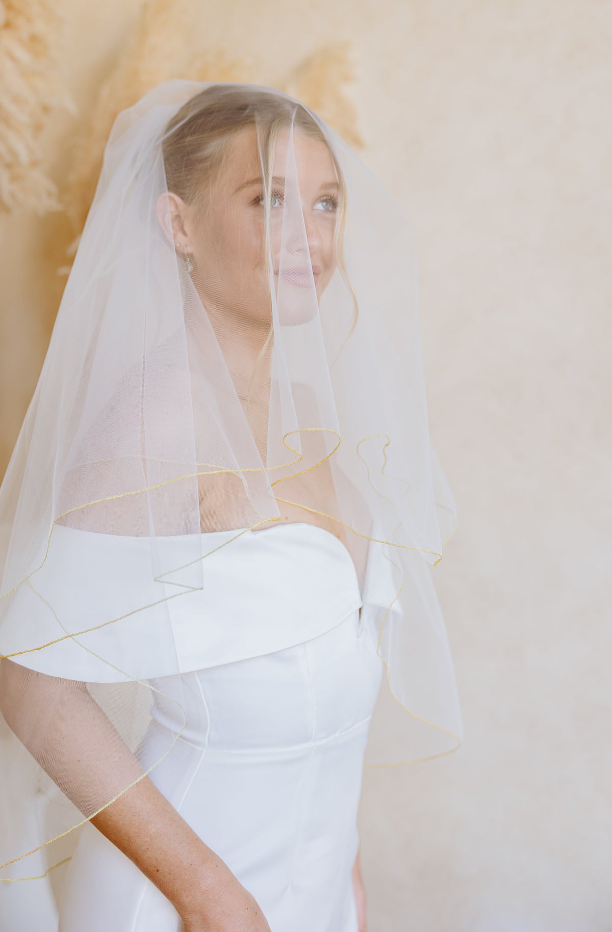 A bride wearing a sheer white tulle bridal veil with delicate gold embroidered accents, paired with a white off-the-shoulder wedding dress, posed against a soft beige background with dried pampas grass.