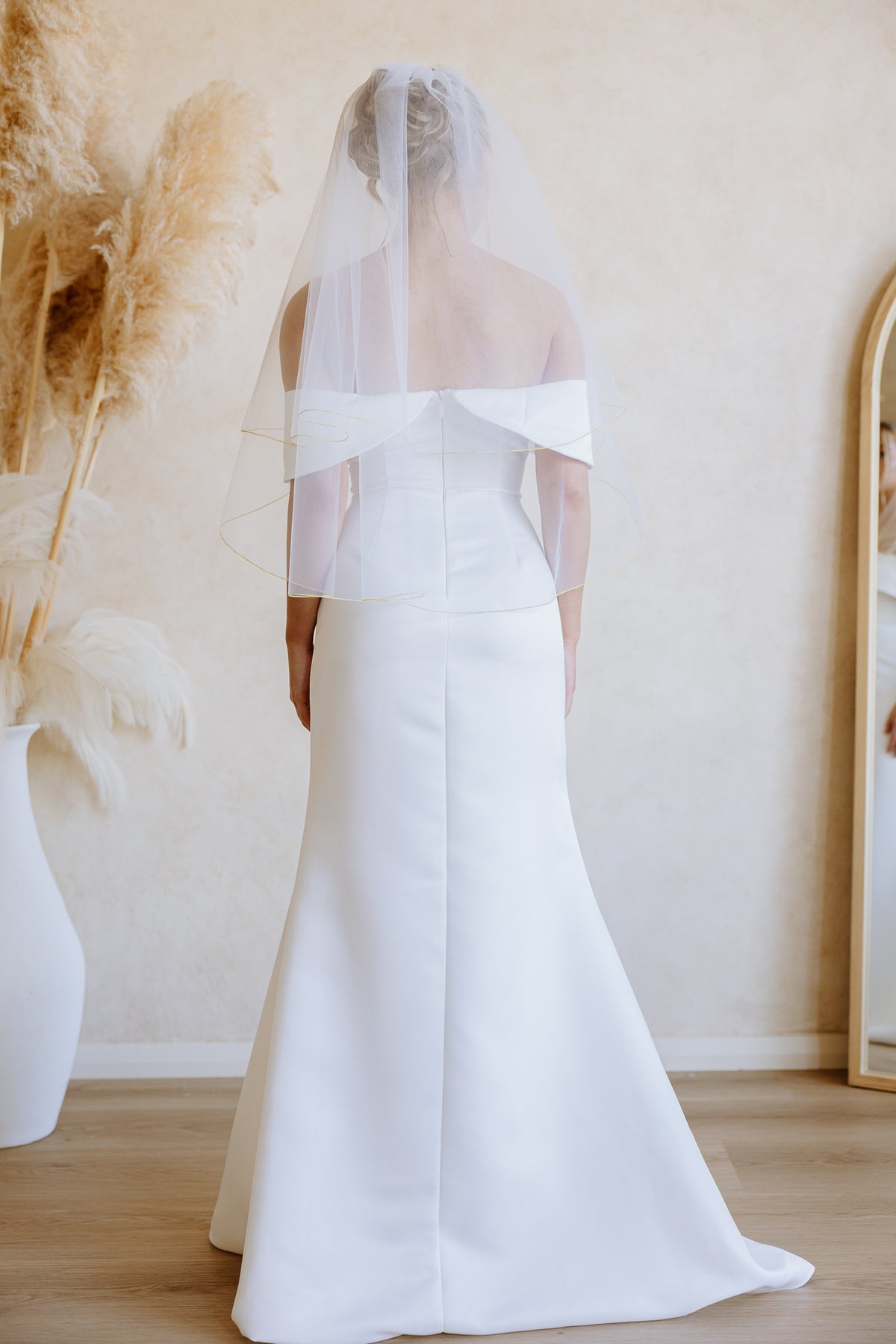 A woman wearing a white off-the-shoulder wedding gown and a long sheer veil, photographed from the back in a softly lit indoor setting with beige walls, wood flooring, and pampas grass decor.