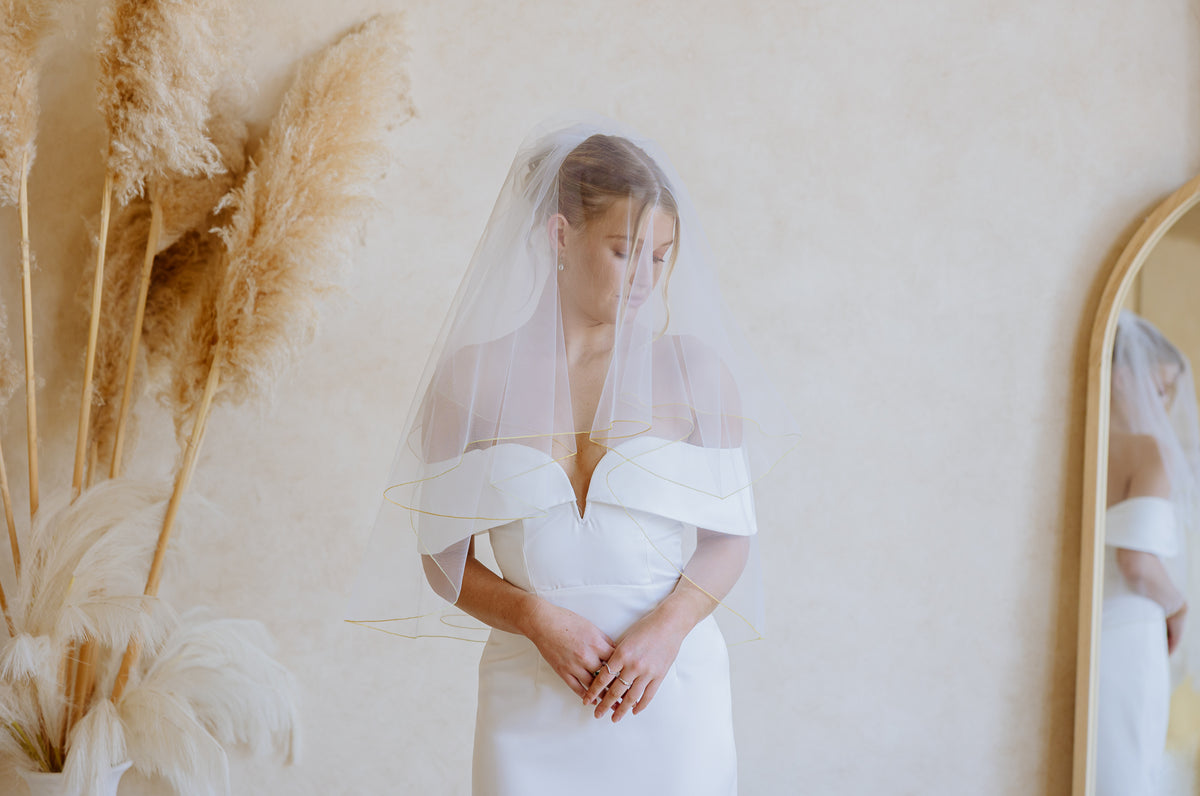 A woman wearing a white off-the-shoulder wedding dress and a sheer white veil stands indoors against a warm beige wall, with dried pampas grass on the left and part of a mirror on the right.