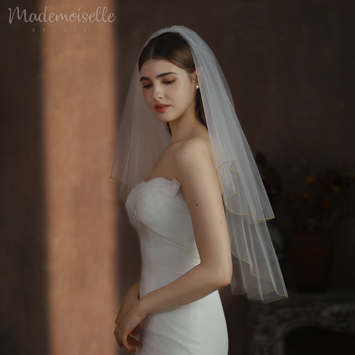 Elegantly draped in a white gown, a bride radiates beauty beneath her exquisite wedding veil with golden threaded trim
