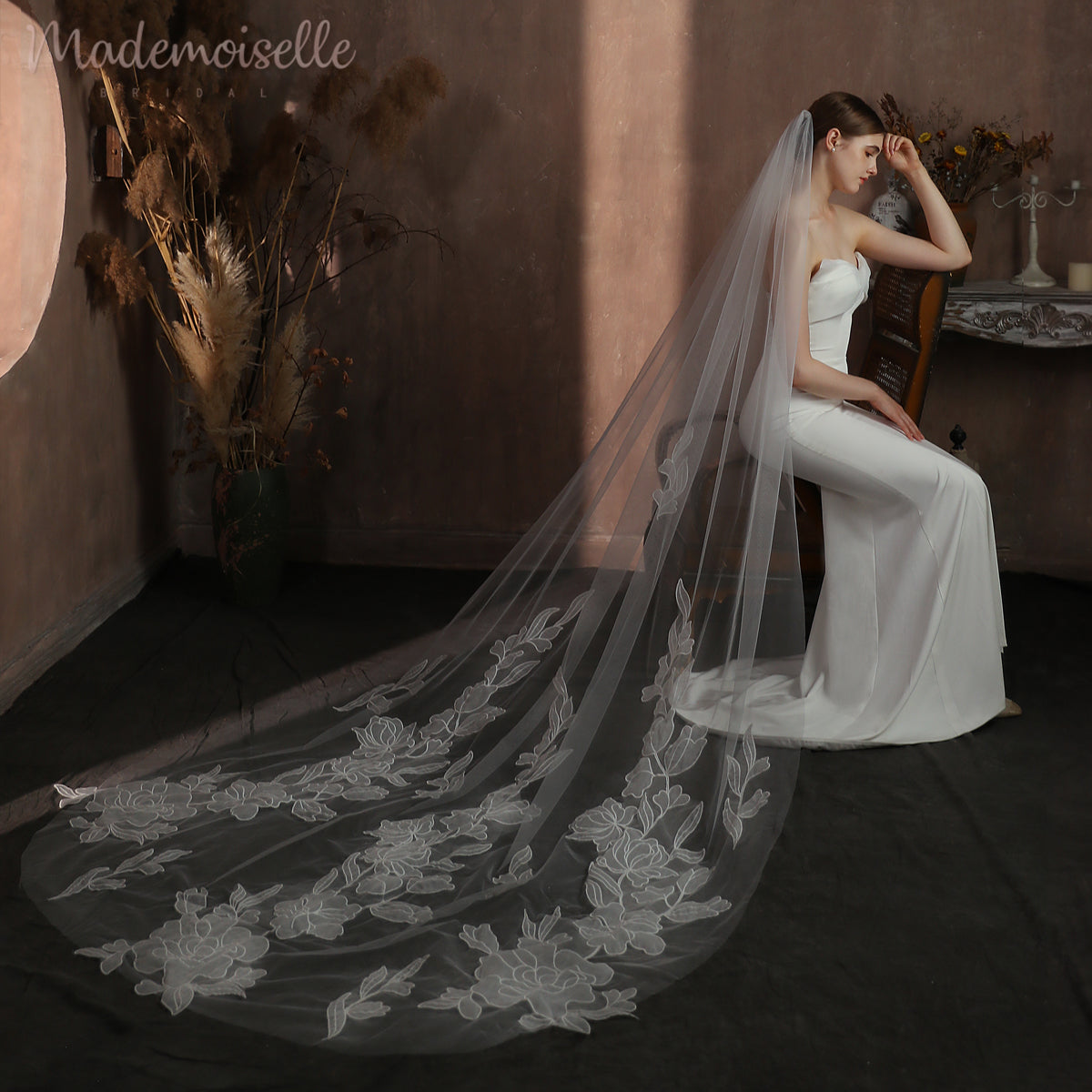The bride, draped in an exquisite veil with floral lace, sits gracefully in a chair, her long veil creating an ethereal aura around her.