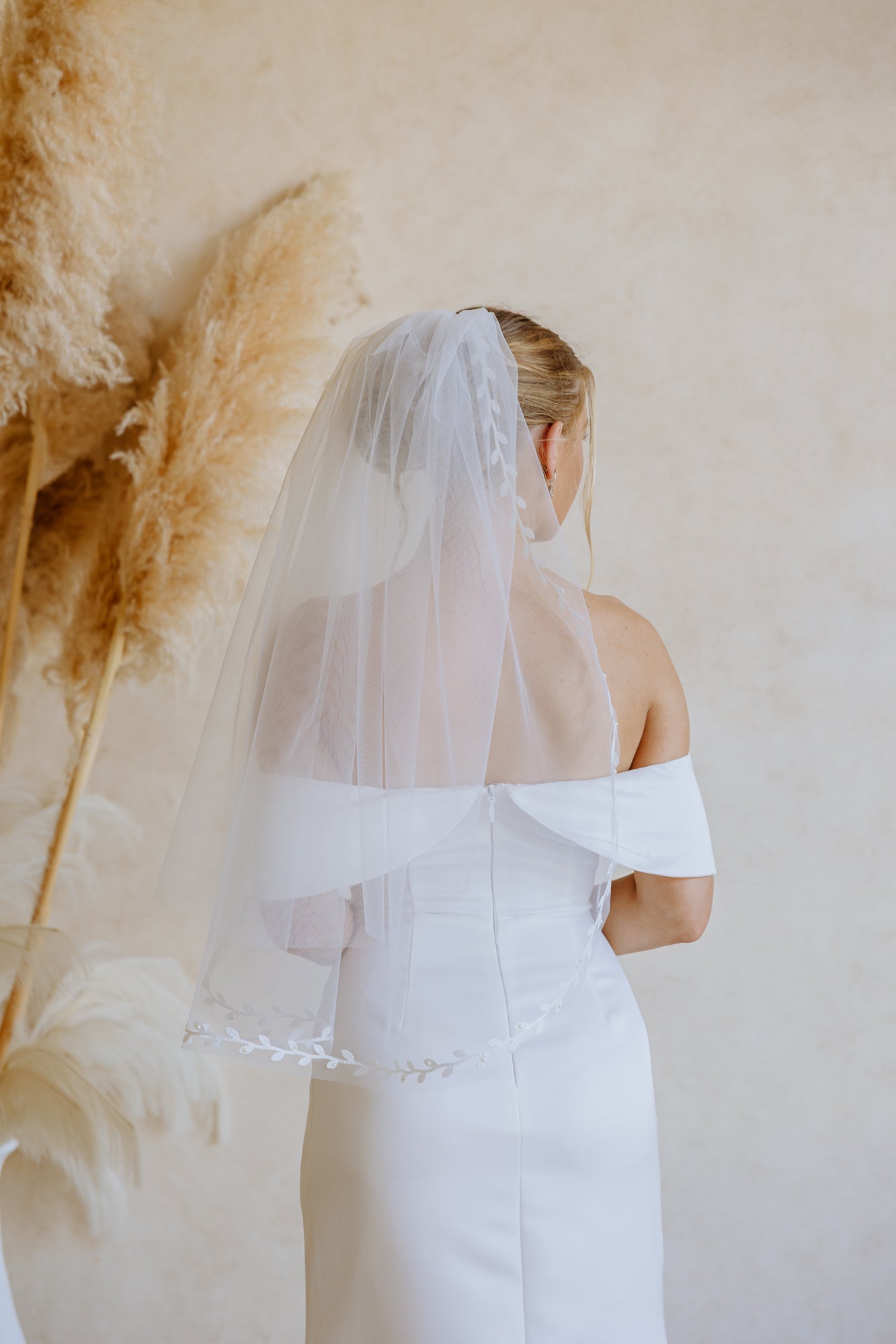 A woman wearing a white off-the-shoulder wedding dress and a sheer white veil with embroidered leaf details, photographed against a soft beige studio background.