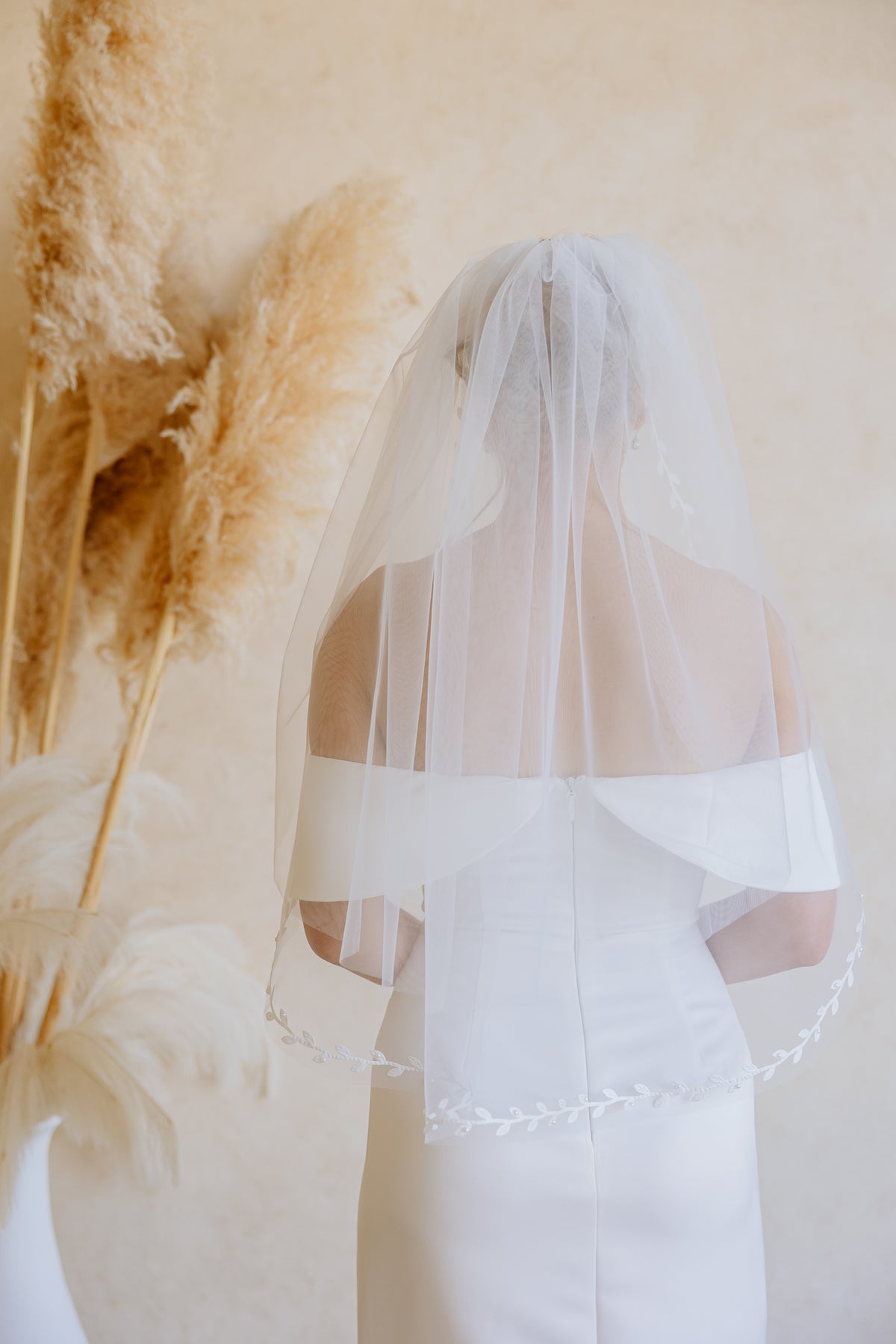 A bride viewed from behind wearing a long white veil over a white wedding dress, standing against a warm beige wall with dried pampas grass decor on the left.