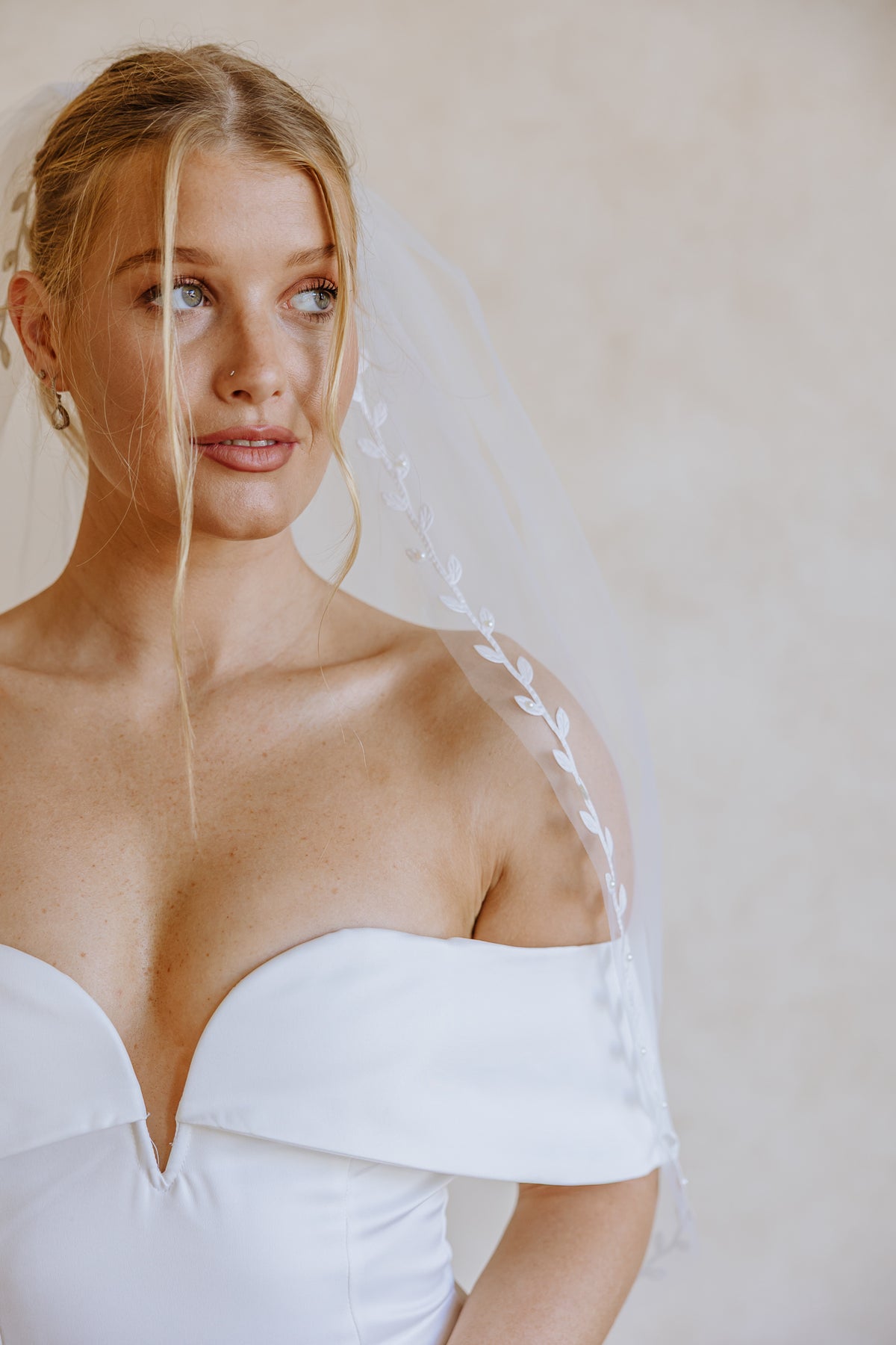 A woman wearing a white off-the-shoulder bridal gown and a sheer white veil with a leafy vine border, photographed against a soft beige wall.