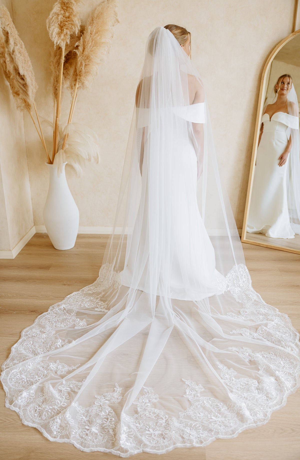 A bride viewed from behind wearing a long white wedding dress with a dramatic lace train and a long sheer veil, standing in a softly lit room with a mirror and pampas grass decor.