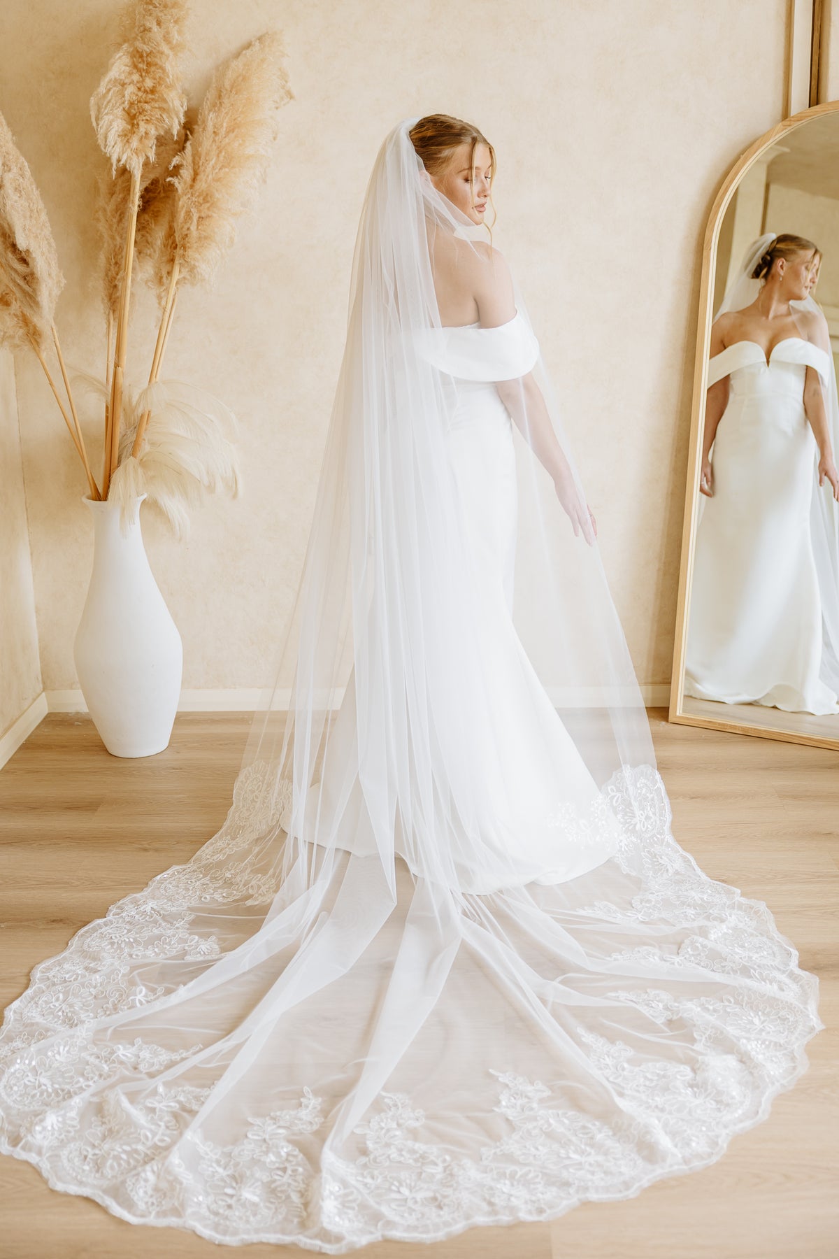 A bride wearing a long white off-the-shoulder wedding dress with a very long sheer veil stands in a softly lit room, with a mirror showing her reflection and dried pampas grass in a white vase beside her.