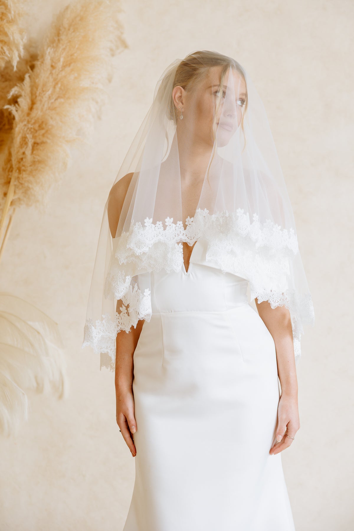 A woman wearing a white wedding dress with a sheer white veil and floral lace trim, standing against a warm beige wall with dried pampas grass on the left.