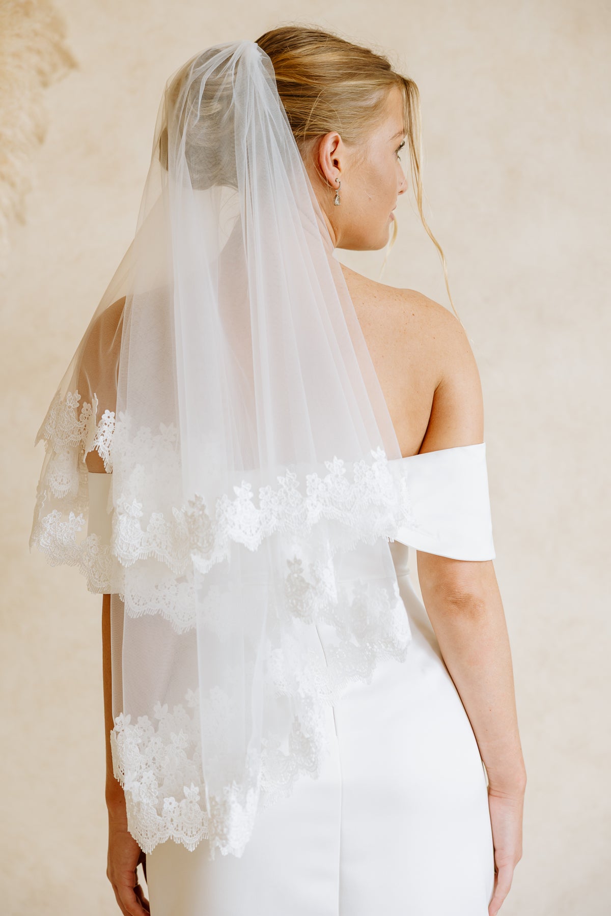 A bride shown from the back wearing a long white tulle veil with layered lace trim over a white off-the-shoulder wedding dress, against a warm beige textured background.