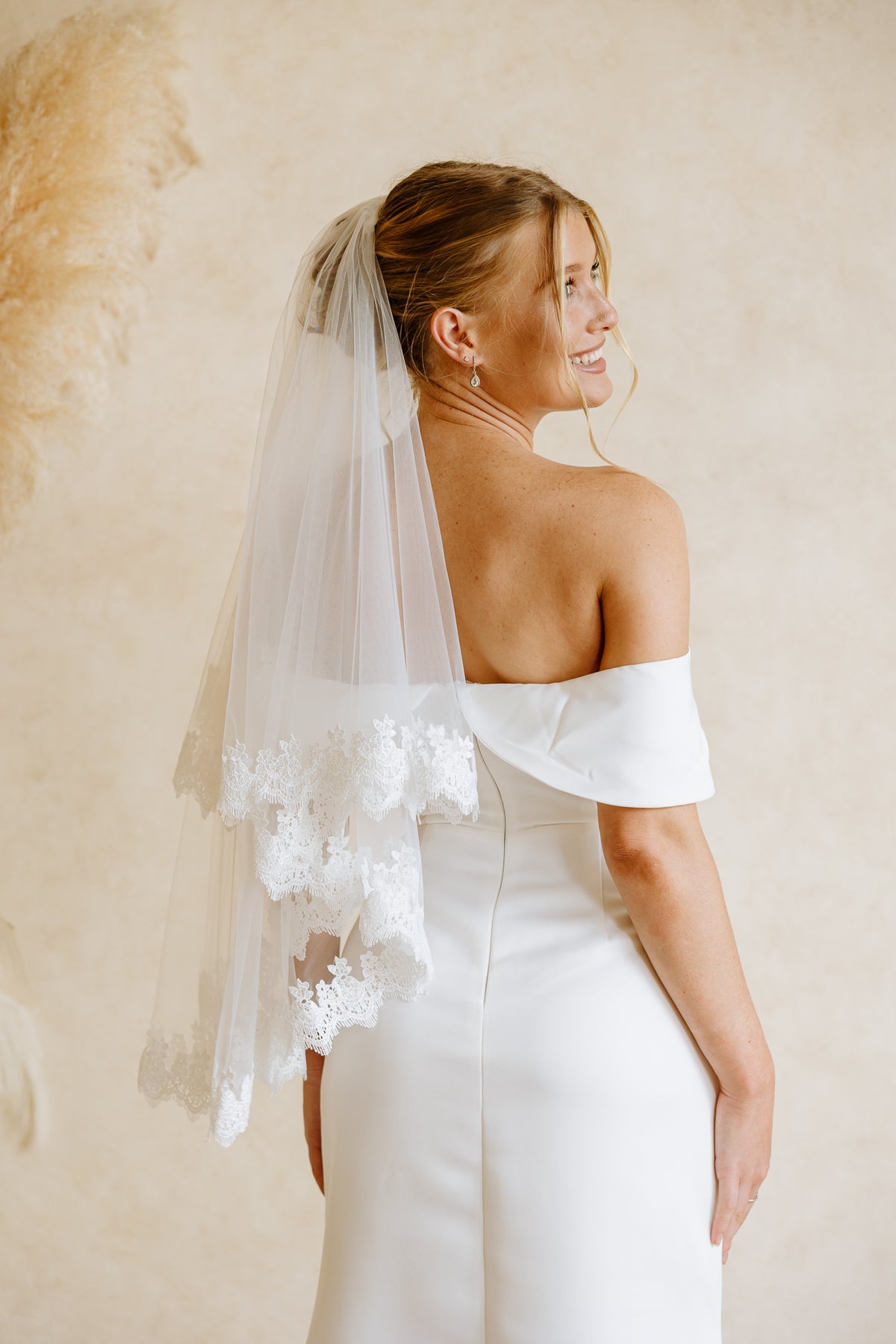 A bride wearing a white off-the-shoulder gown and a long layered tulle veil with floral lace trim, photographed from the back against a warm beige wall.
