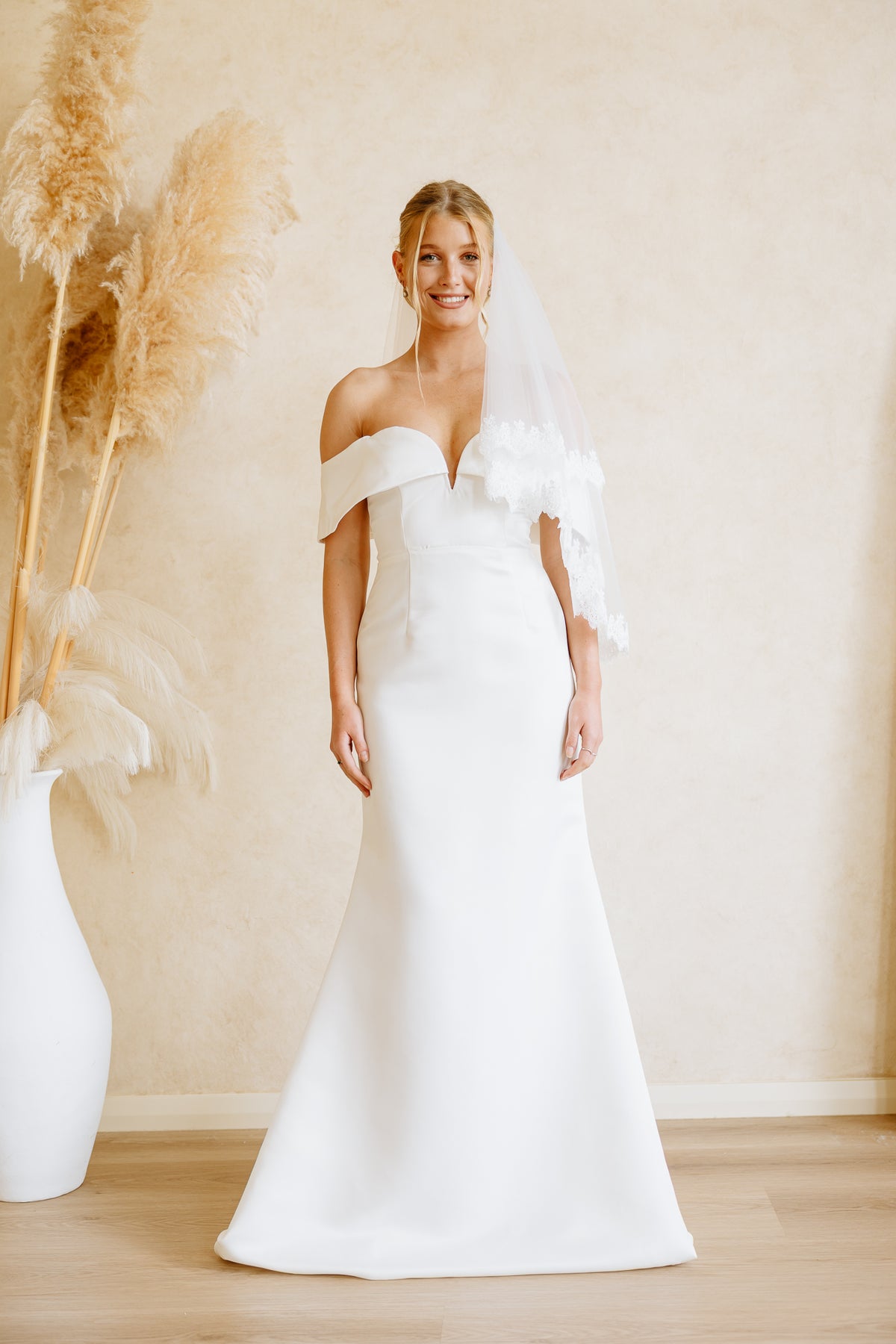 A woman wearing a white off-the-shoulder wedding dress with a matching sheer veil stands in a softly lit room with a beige wall and light wood floor. Decorative pampas grass in a white vase is visible on the left.