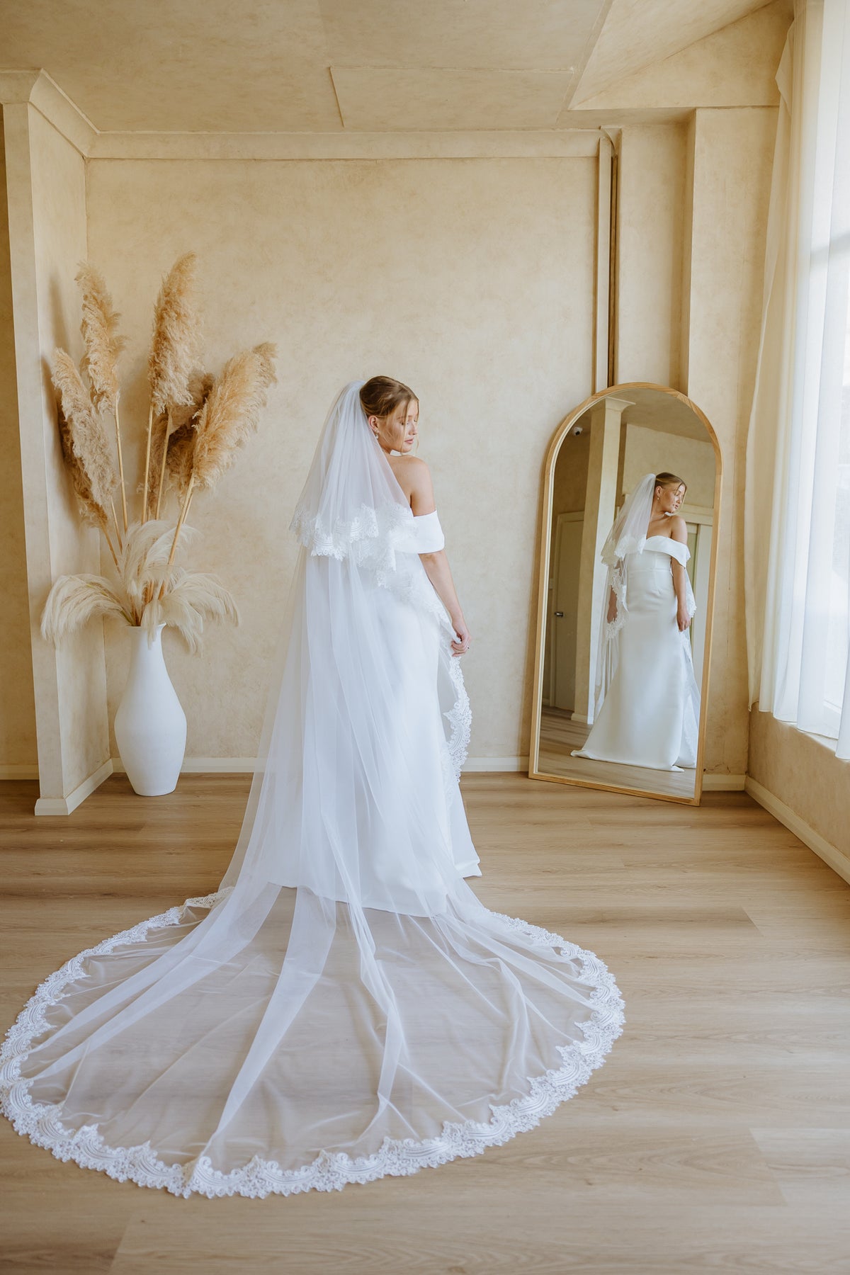 A bride in a white off-the-shoulder wedding dress with a long sheer tulle train and veil stands in a warm beige room, looking into a gold-framed mirror that reflects her profile. A white vase with dried pampas grass sits to the left.