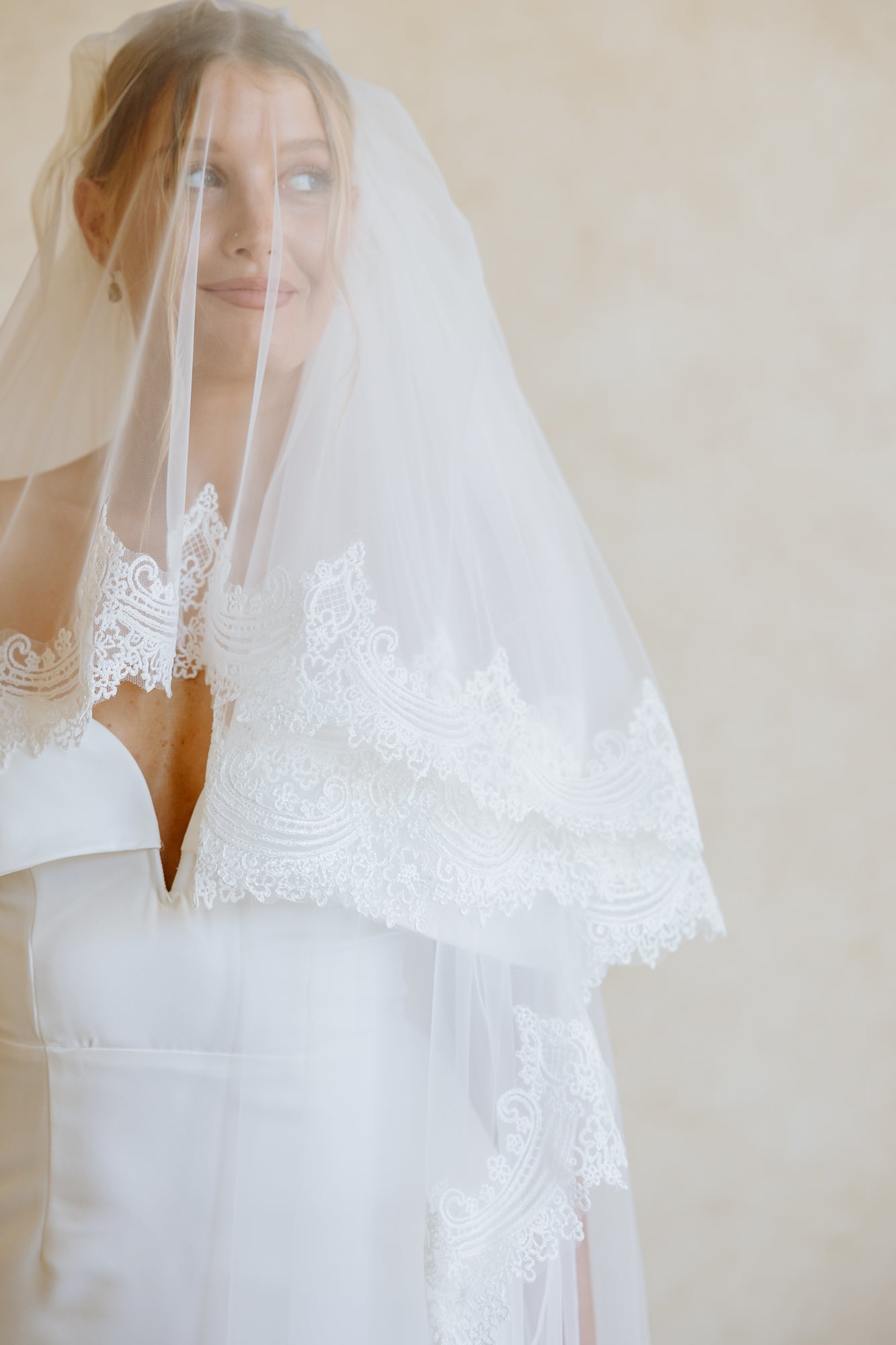 A bride in a white wedding gown and long sheer veil stands with her back to the camera in a warm beige room, with a large mirror showing her front reflection and dried grass decor in a white vase.