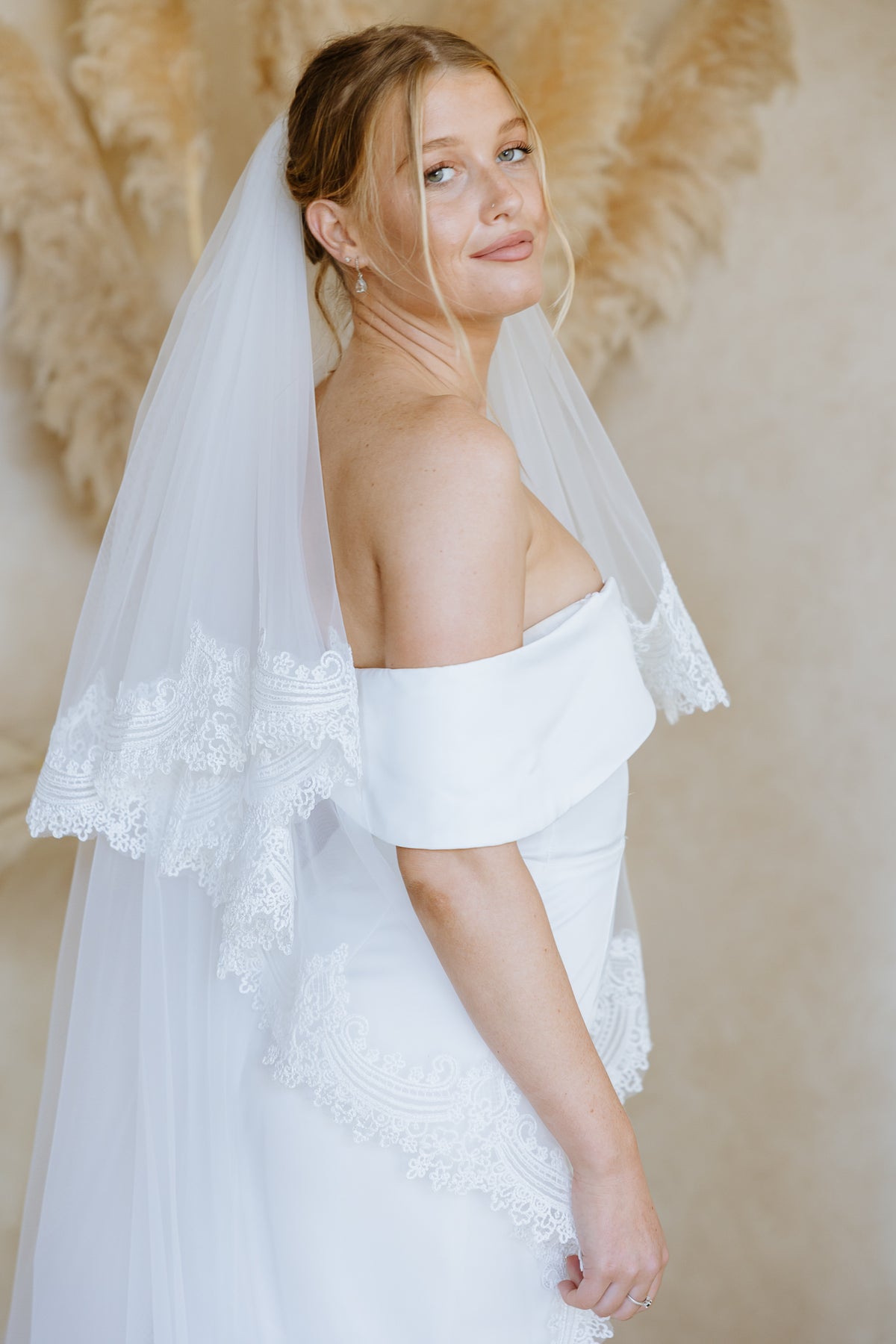 A woman wearing a white off-the-shoulder wedding dress with lace trim and a long sheer veil, posed against a warm beige background with soft decorative foliage.