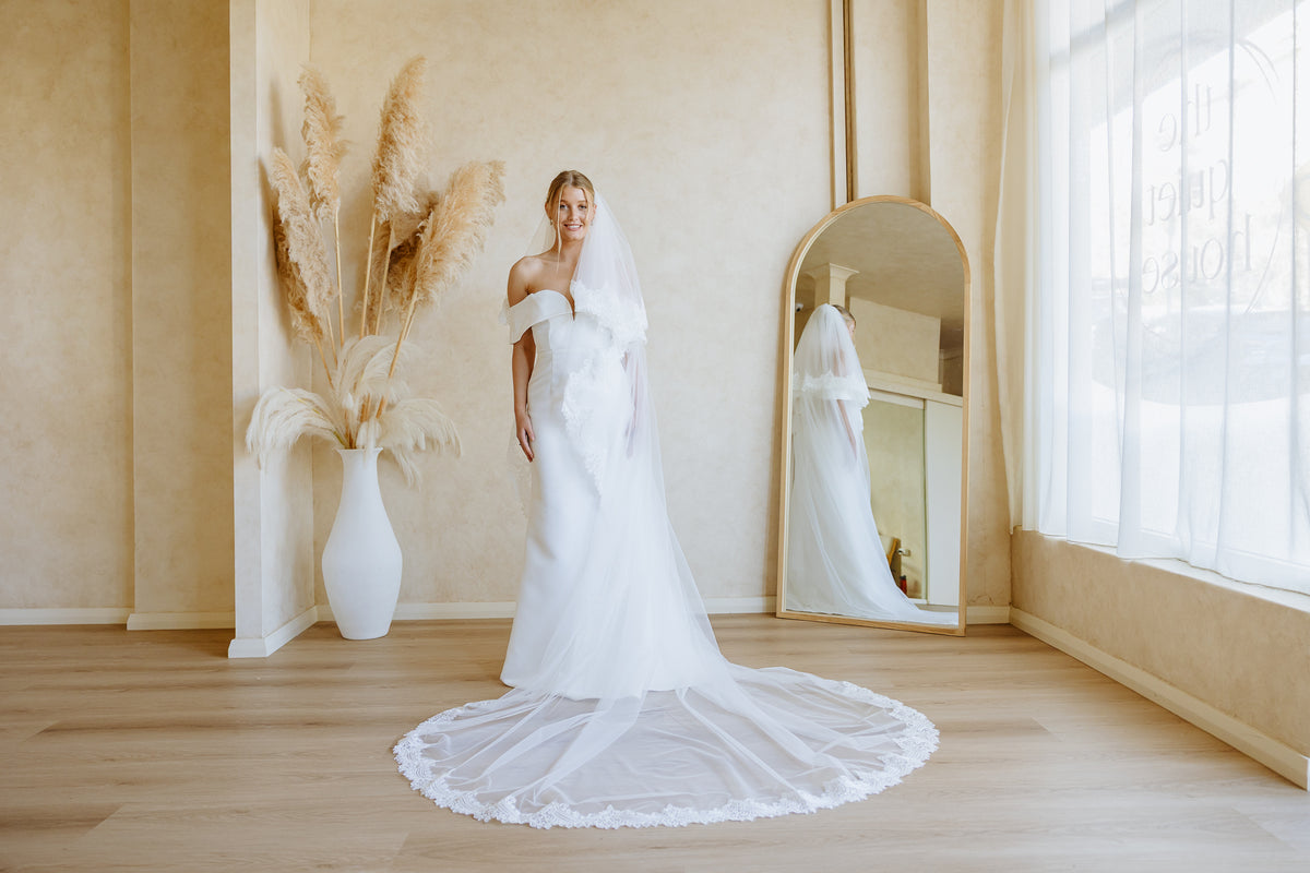 A woman wearing a white off-the-shoulder wedding gown with a long sheer veil stands in a bright room with wood floors, a large vase with pampas grass, and a tall arched mirror reflecting her profile.