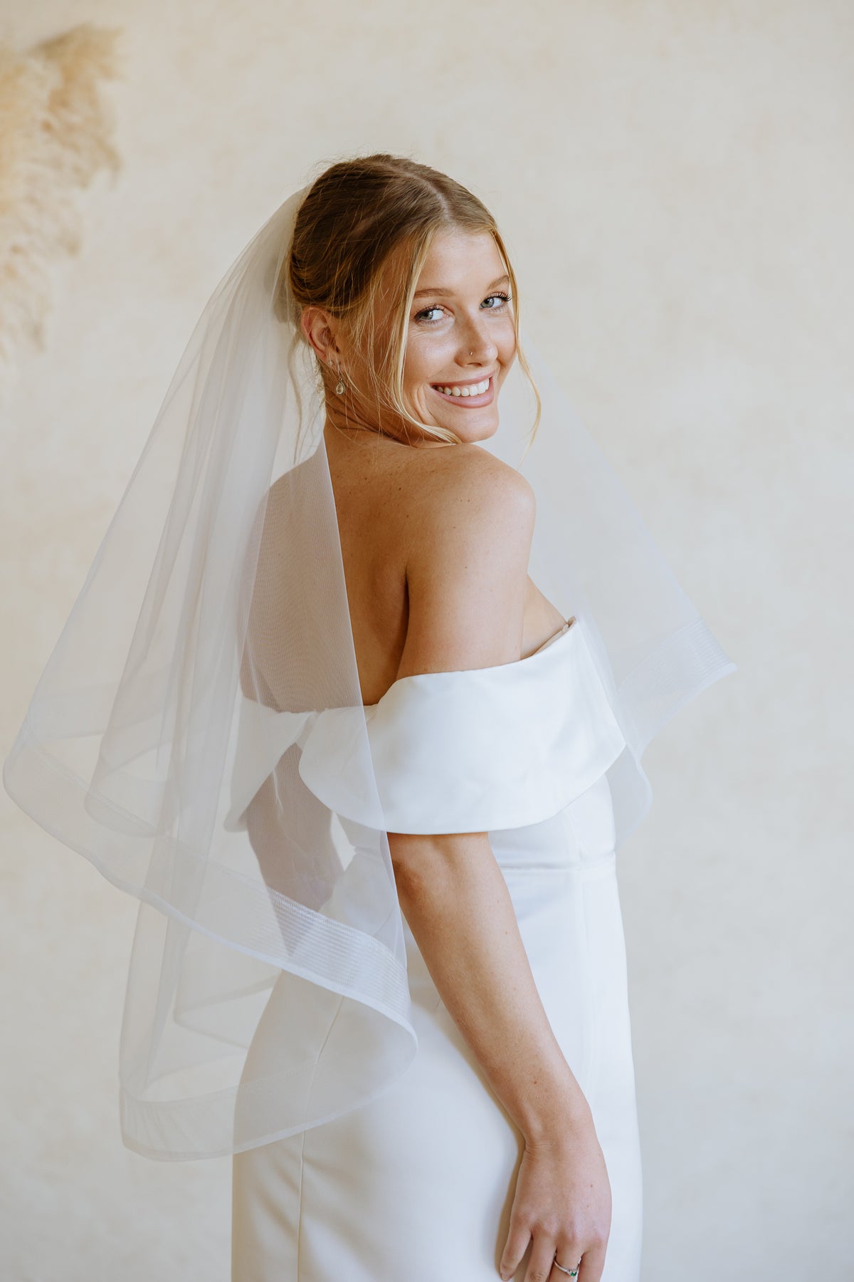 A smiling bride wearing an off-the-shoulder white wedding dress and a sheer veil with horsehair trim, photographed against a soft beige textured wall.